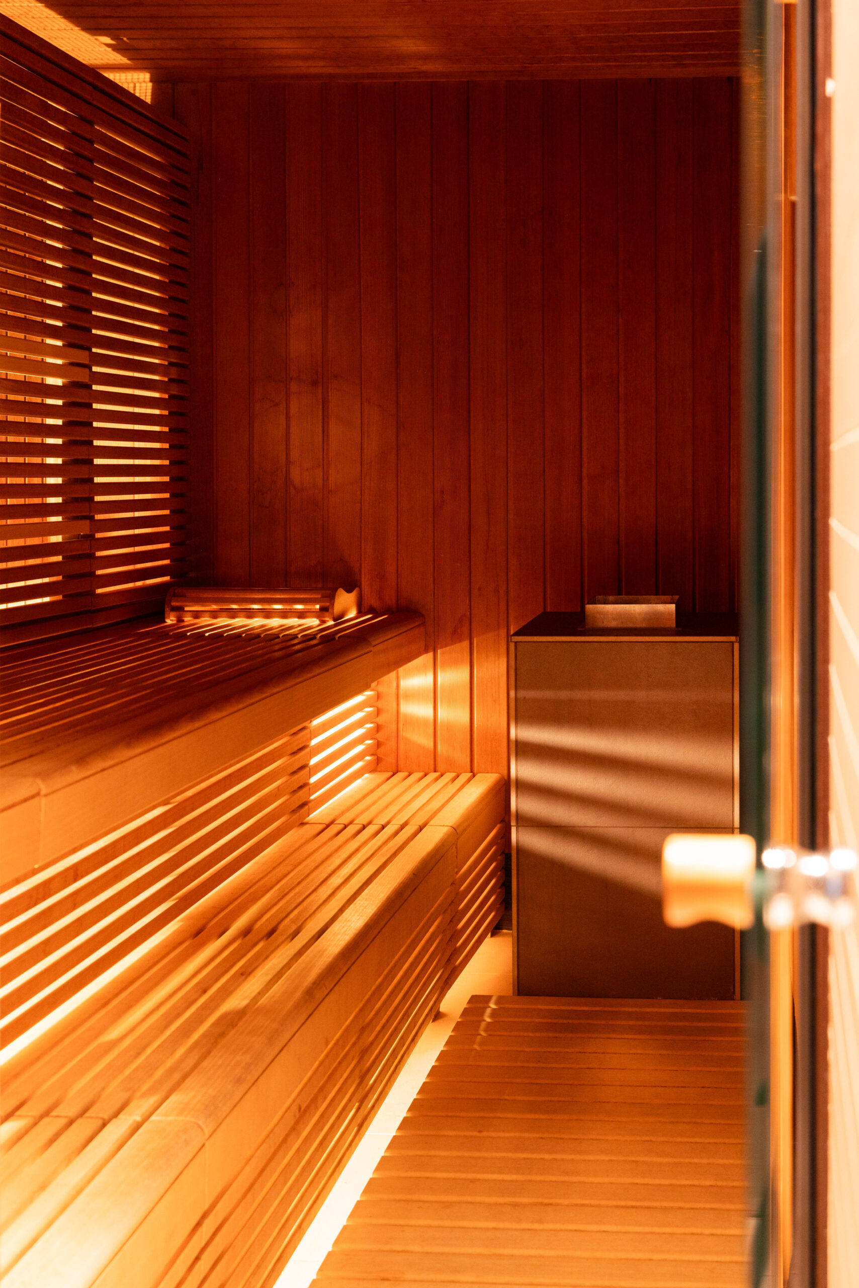 Eine warme, hölzerne Sauna im Sauna Spa Hotel Sonne mit Lattenbänken an den Wänden, sanfter Beleuchtung und einem Ofen in der Ecke - vom Eingang aus gesehen mit einem auf der Bank gefalteten Handtuch.
