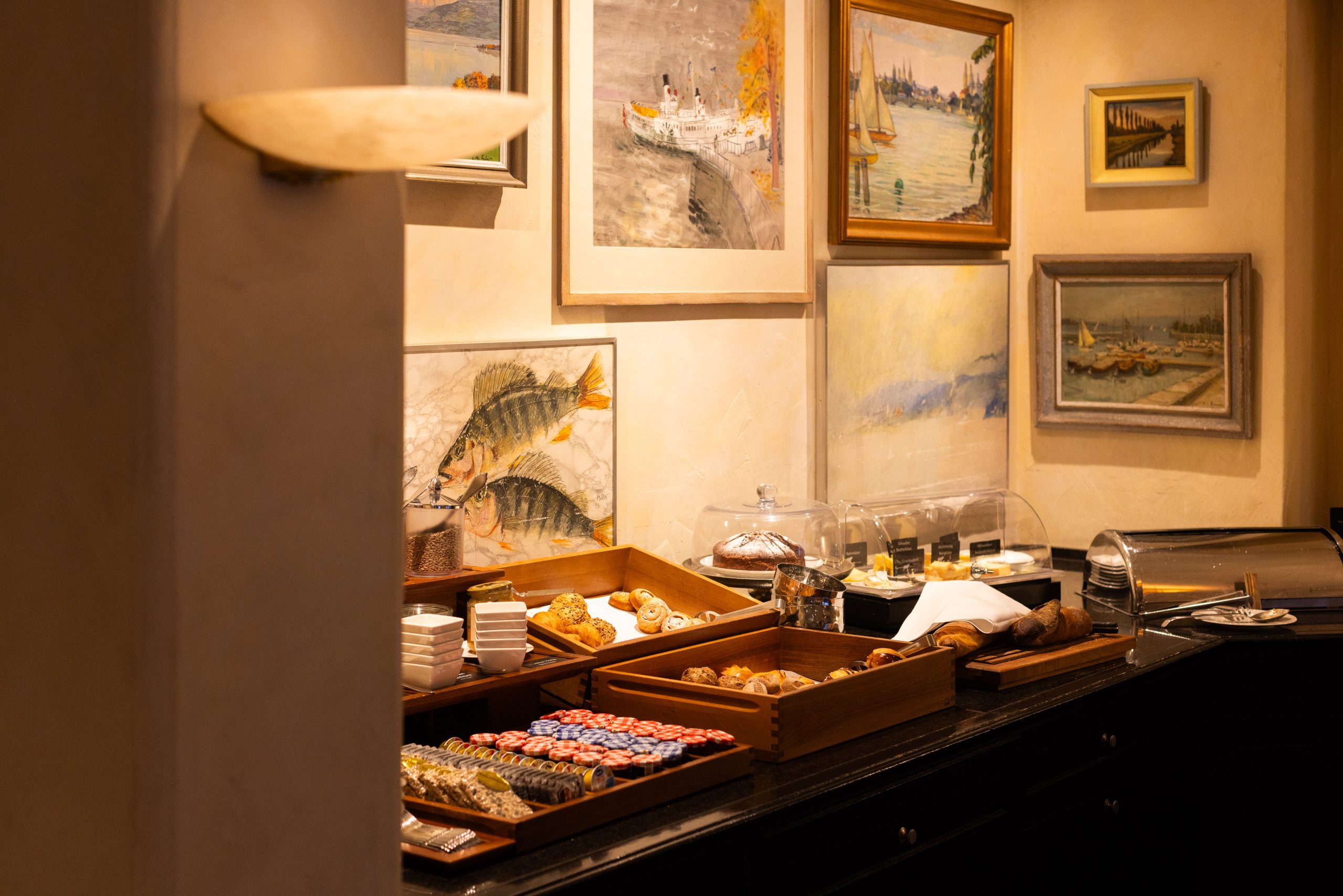 Ein Frühstücksbuffet mit Tabletts mit Gebäck, Butter- und Marmeladenpaketen, Tellern, Utensilien und Abdeckungen, die an einer mit gerahmten Bildern von Booten, Fischen und Landschaften geschmückten Wand stehen.