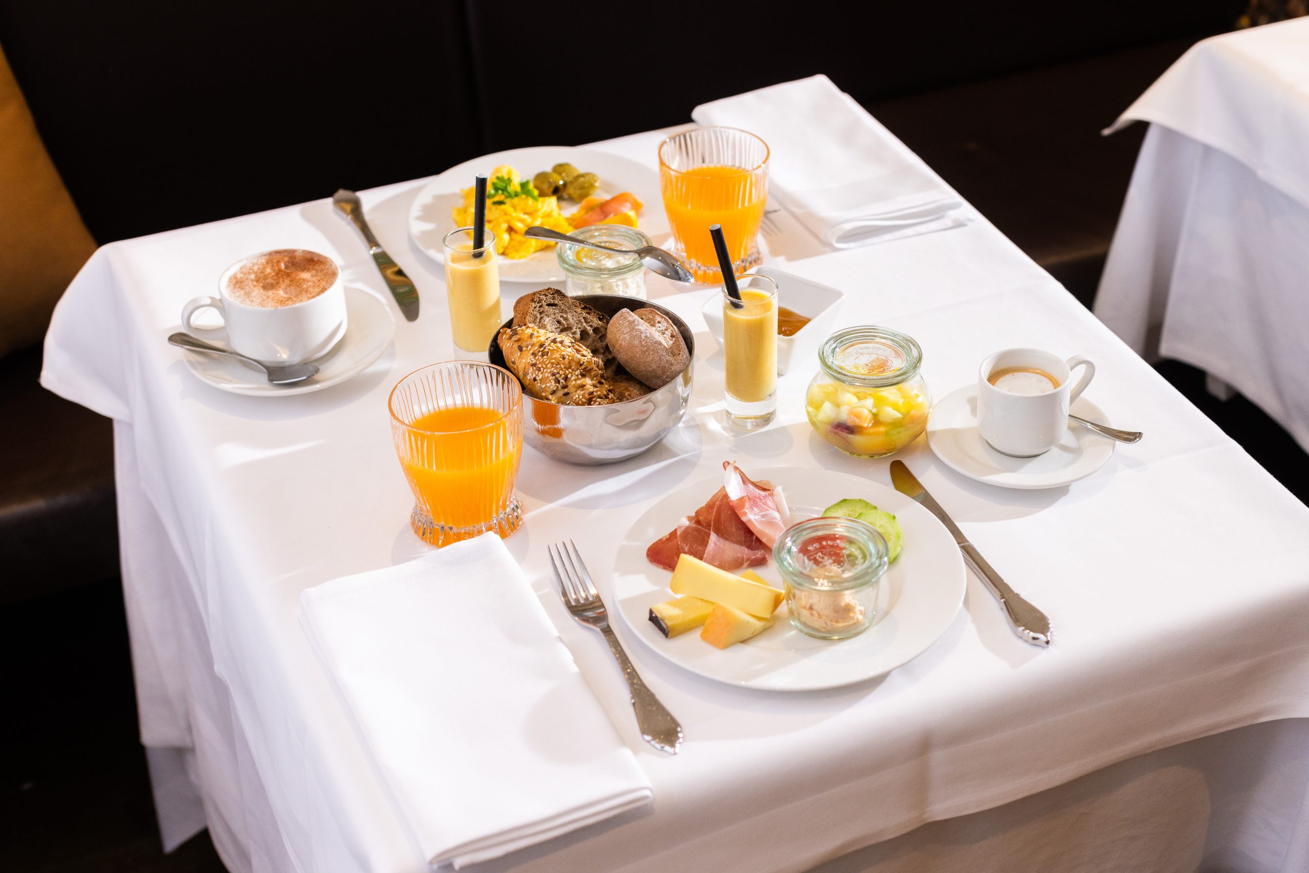 Ein gedeckter Tisch für zwei Personen mit verschiedenen Brotsorten, Aufschnitt, Käse, geschnittenem Obst, Gläsern mit Orangensaft, Smoothies, Cappuccino und Espresso auf weißen Tischtüchern - perfekt für ein entspanntes Frühstück.