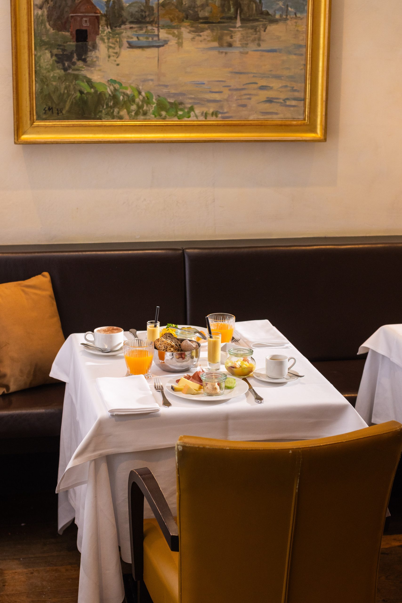 Ein kleiner Esstisch für das Frühstück mit Kaffee, Saft, Gebäck, Obst und Joghurt steht in einem gemütlichen Restaurant mit einer braunen Sitzbank, einem gelben Stuhl und einem gerahmten Gemälde an der Wand.