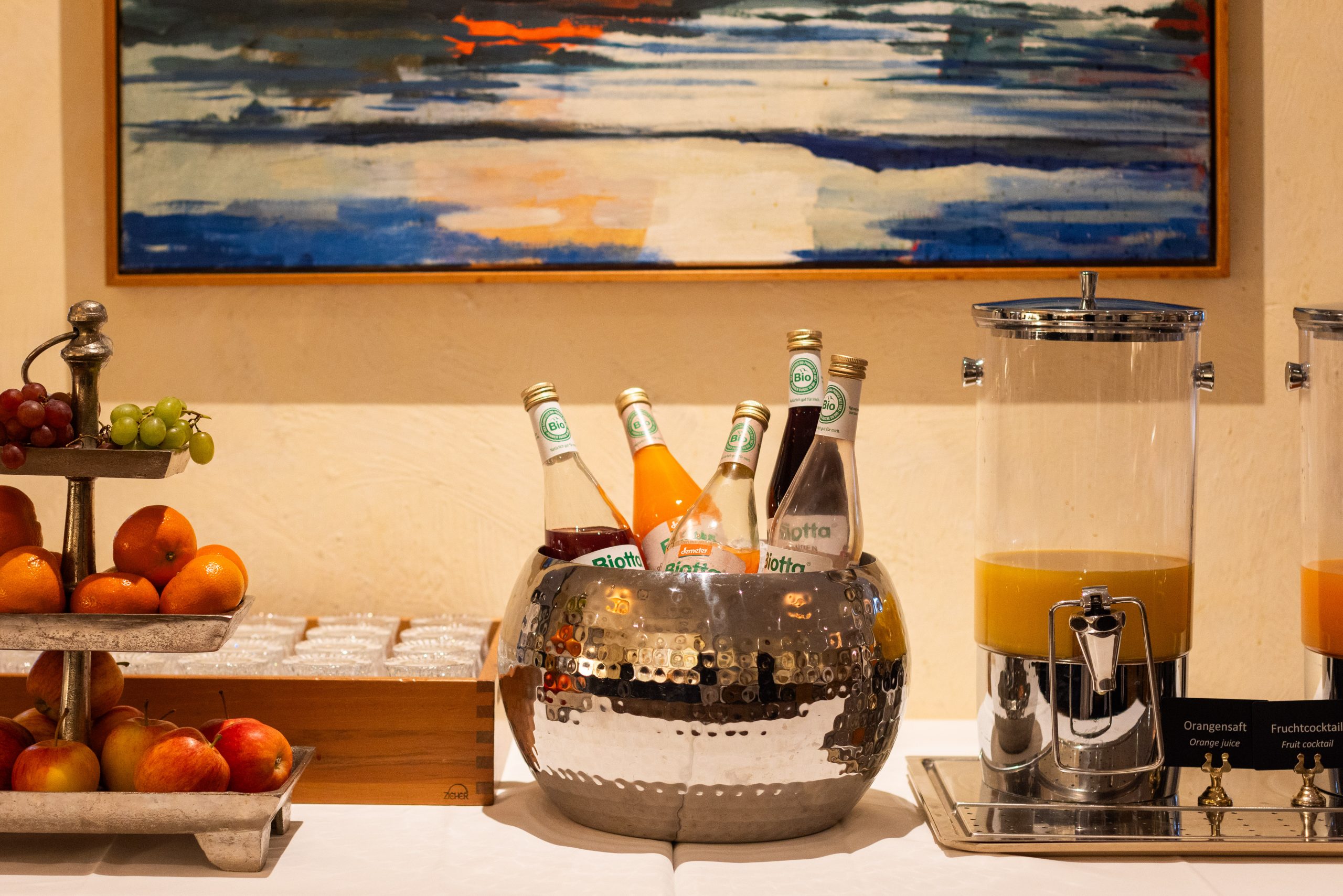 Ein für das Frühstück gedeckter Buffet-Tisch mit einem Obststand mit Äpfeln und Weintrauben, einer Metallschüssel mit Getränkeflaschen und einem Saftspender. An der Wand im Hintergrund hängt ein buntes Gemälde.