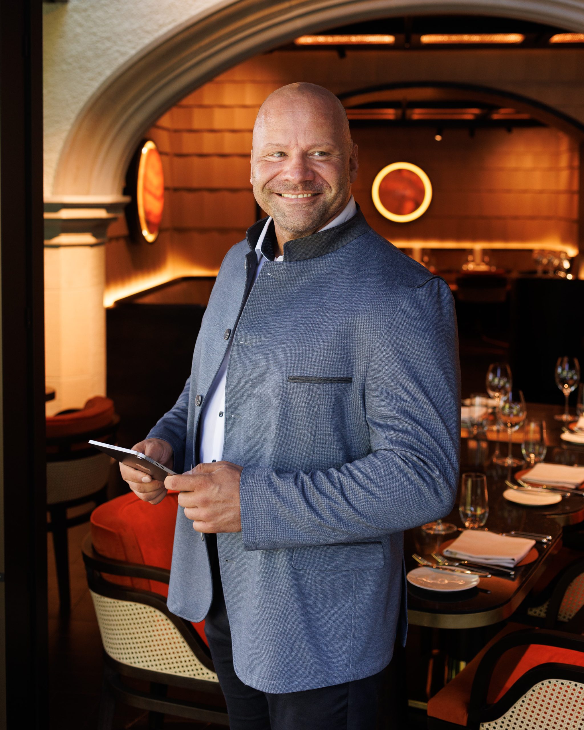 Ein lächelnder Mann in einer blauen Jacke steht in einem stilvollen Restaurant mit gedeckten Tischen und warmer Beleuchtung und hält ein Tablet in der Hand.