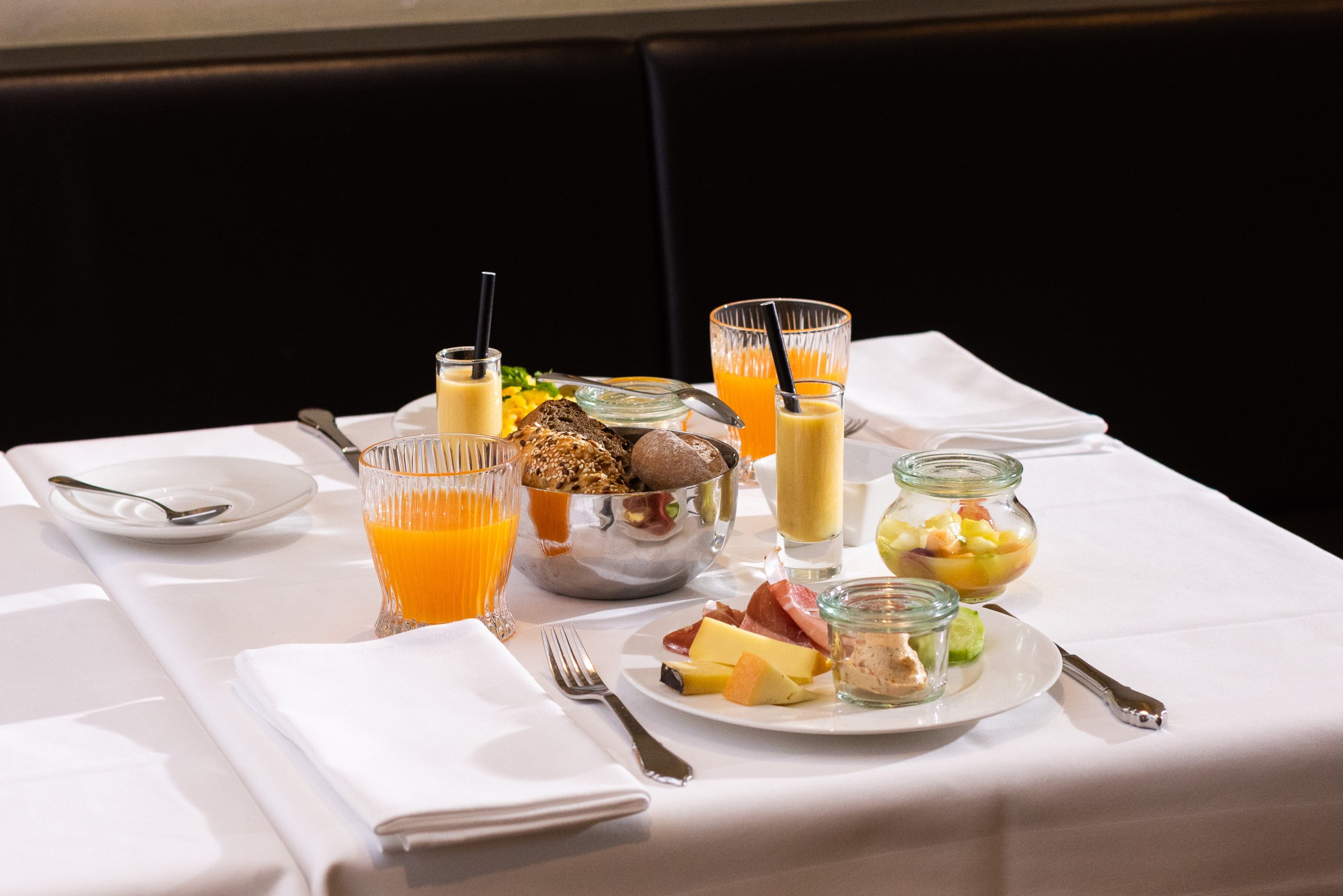Auf einem ordentlich gedeckten Tisch für zwei Personen im Frühstück Boutiquehotel Sonne stehen Orangensaft, kleine Gläser mit Obstsalat, Teller mit verschiedenen Käsesorten, Wurstwaren, frisches Brot und eine Schale mit verschiedenen Brötchen auf einer weißen Tischdecke.
