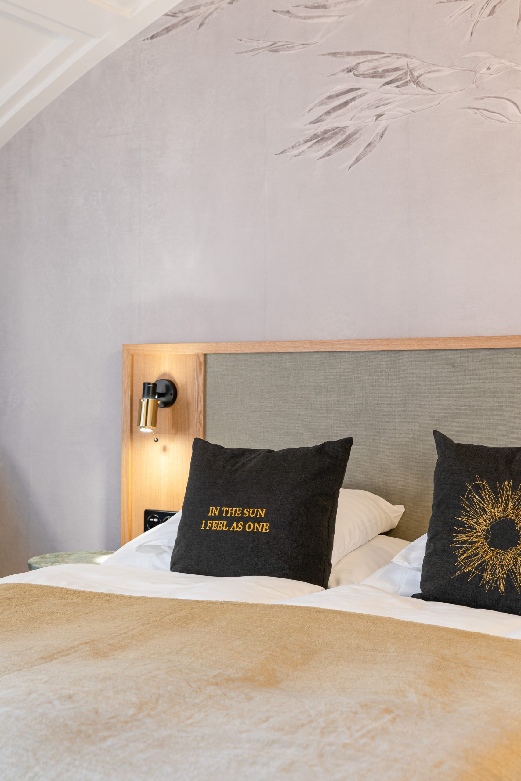 Ein sauber gemachtes Hotelzimmerbett mit zwei dunklen Zierkissen - eines mit Sonnenschliffmuster, das andere mit der Aufschrift IN THE SUN I FEEL AS ONE - steht vor einer hell gestrichenen Wand und einem hölzernen Kopfteil, das die von der Sonne inspirierte Wärme ausstrahlt.