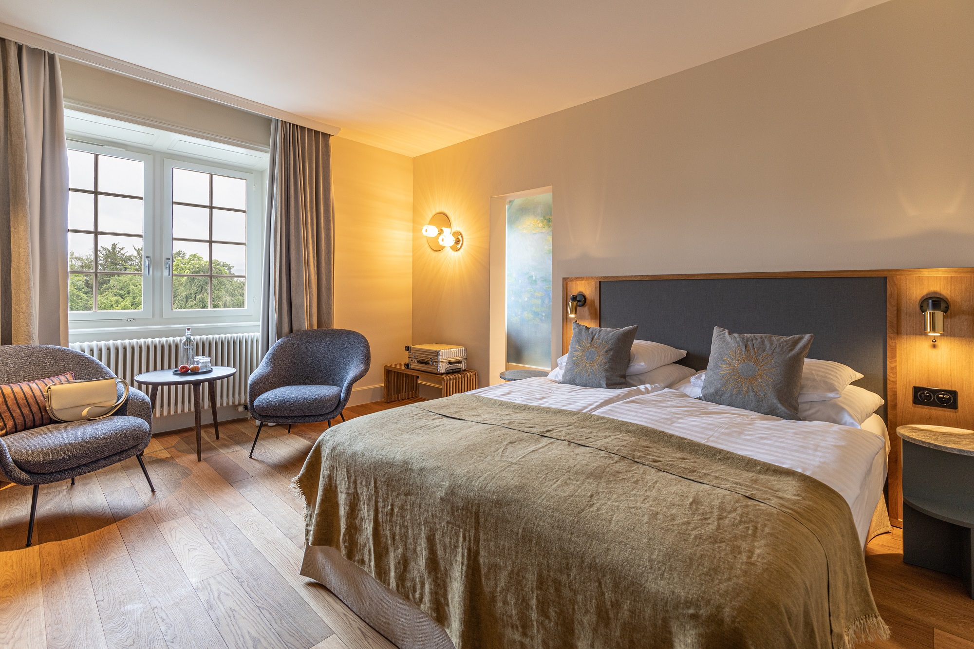 Ein gemütliches Boutiquehotelzimmer mit einem großen Bett, zwei blauen Stühlen und einem kleinen Tisch am Fenster, Holzböden, warmer Beleuchtung und neutralem Dekor.
