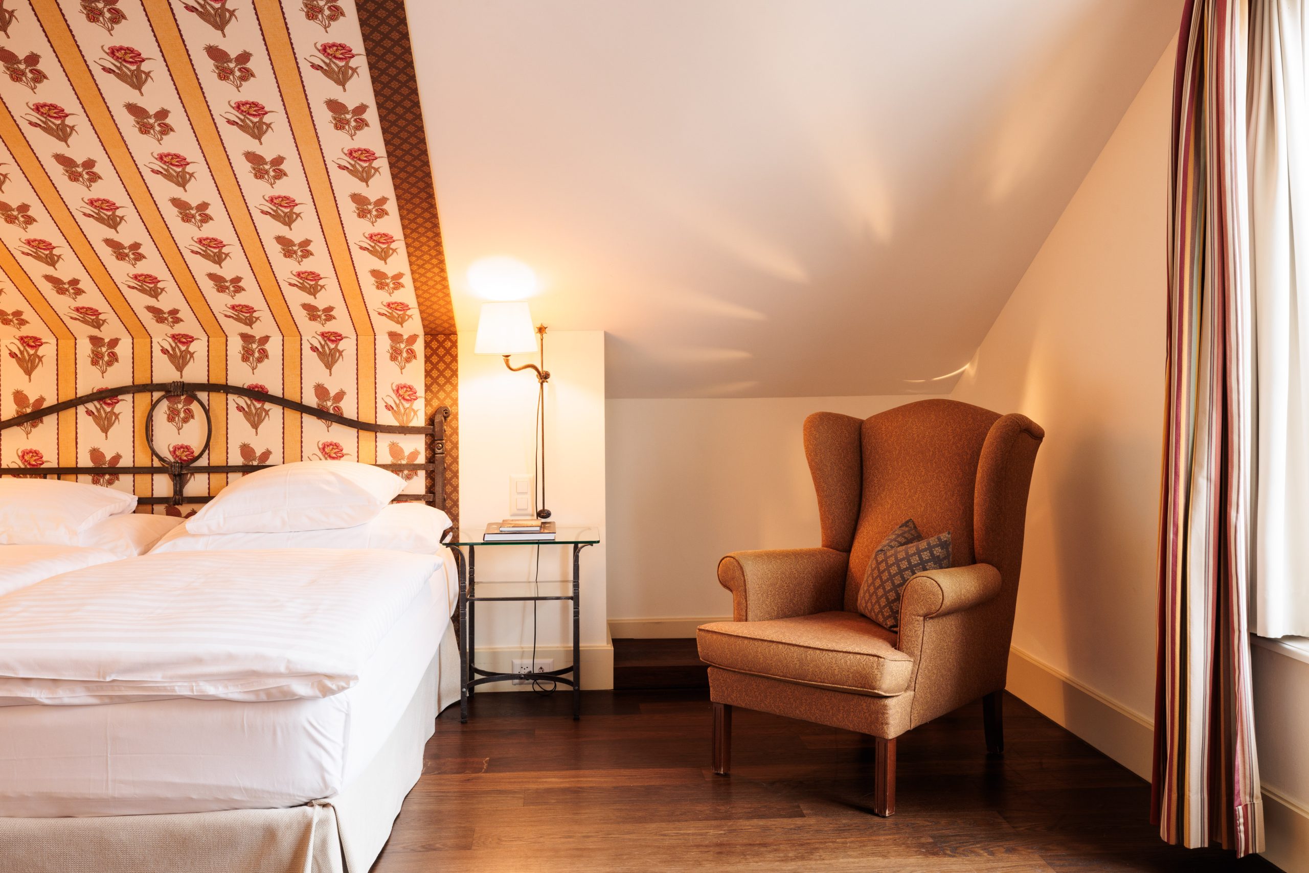 Ein gemütliches Boutiquehotel-Schlafzimmer mit einem Doppelbett, einem gestreiften Ohrensessel, einem Beistelltisch mit einer Lampe, gemusterter Tapete an einer schrägen Wand und einem Fenster mit gestreiften Vorhängen. Das Zimmer hat einen warmen Holzfußboden.