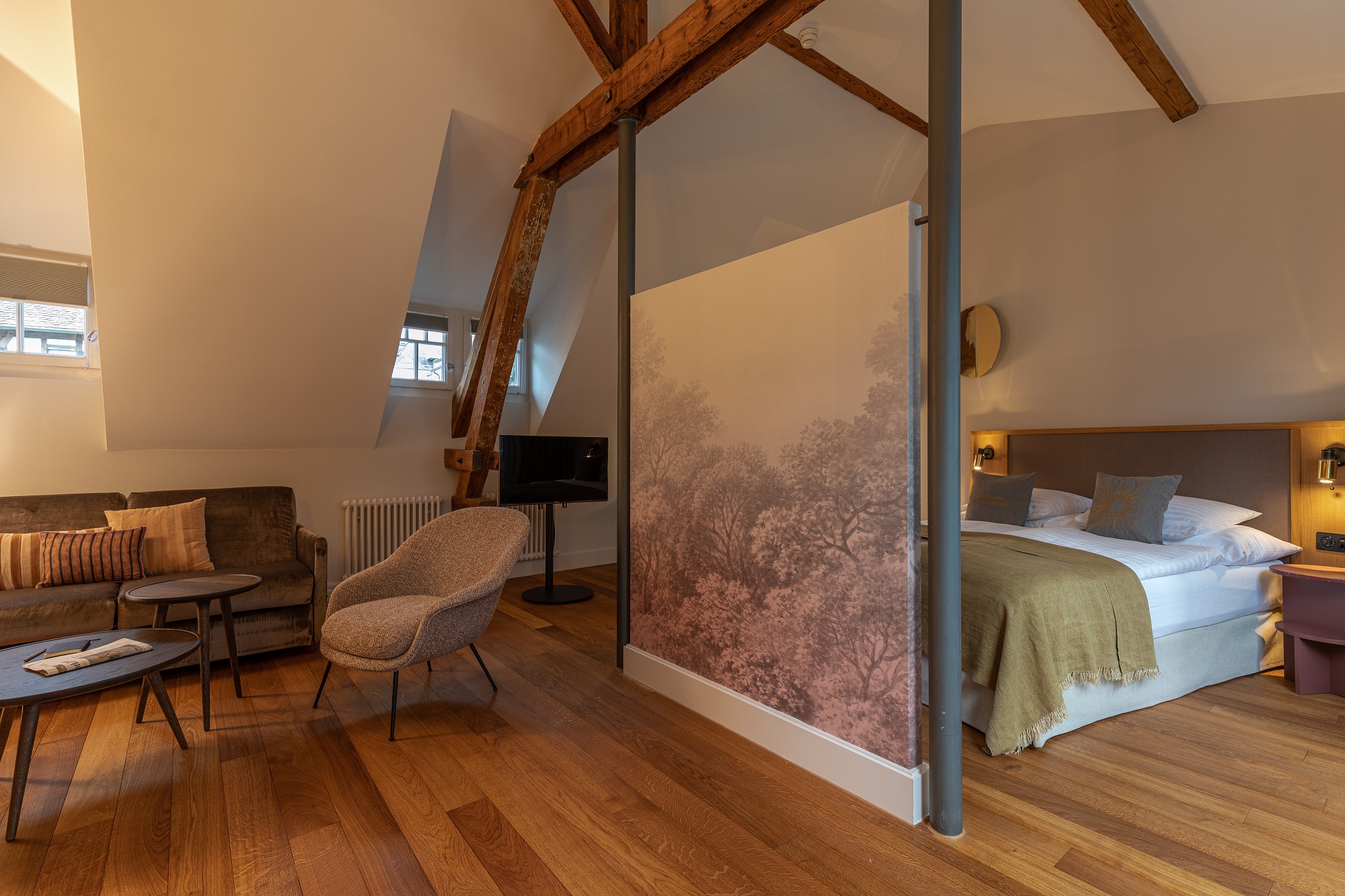 Ein gemütliches Boutique-Hotelzimmer mit Holzboden, freiliegenden Holzbalken, einem Doppelbett mit grünen Akzenten, einem Sofa, Sessel, kleinen Tischen und einem dekorativen Paravent mit Baummotiv, der den Schlafbereich teilt.