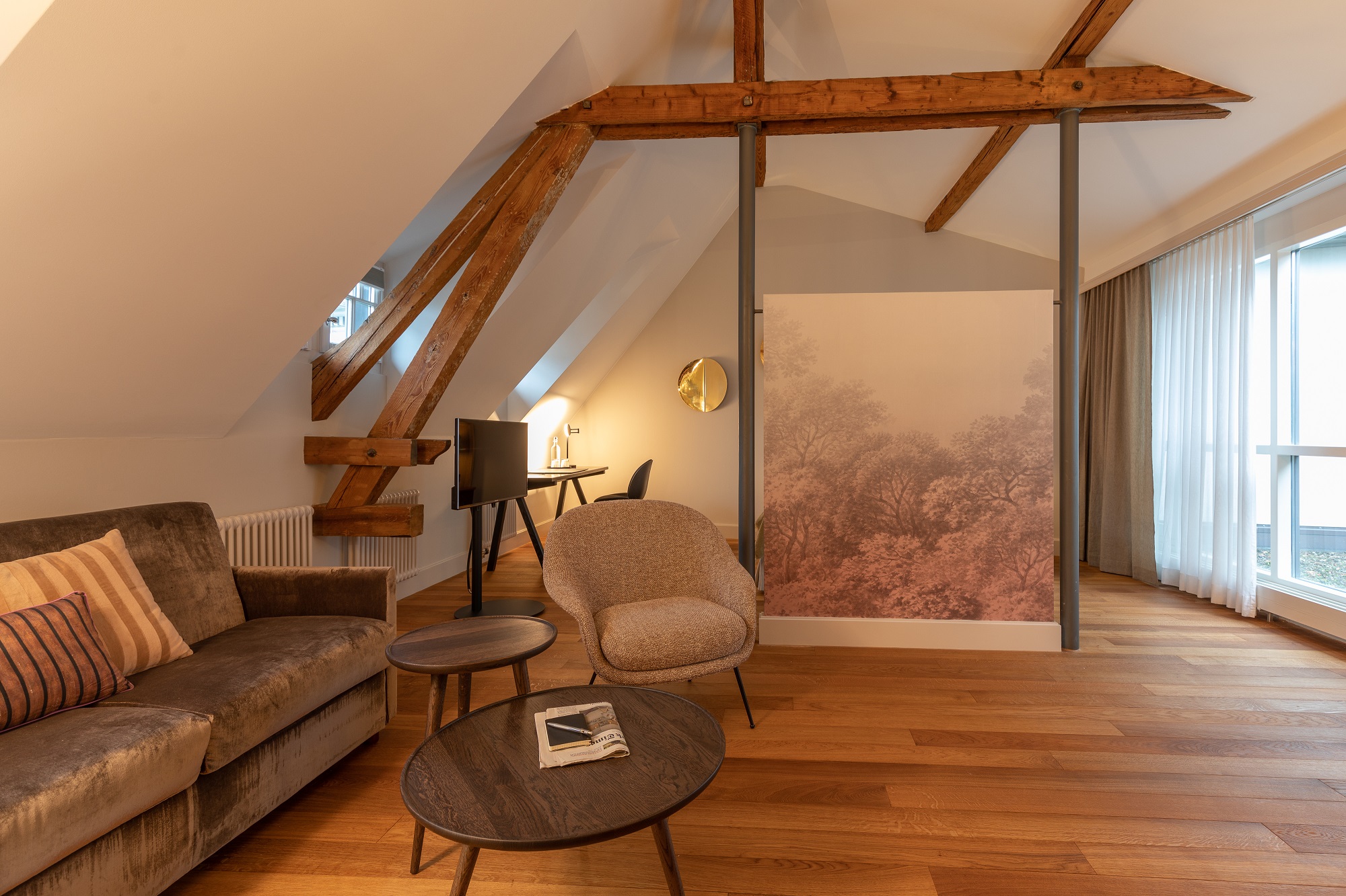 Ein moderner Loft-Wohnbereich im Boutiquehotel-Stil mit freiliegenden Holzbalken, einem grauen Sofa, einem Sessel, runden Couchtischen aus Holz, einem Fernseher, einem Schreibtisch mit Stuhl und großen Fenstern mit transparenten Vorhängen, die natürliches Licht hereinlassen.