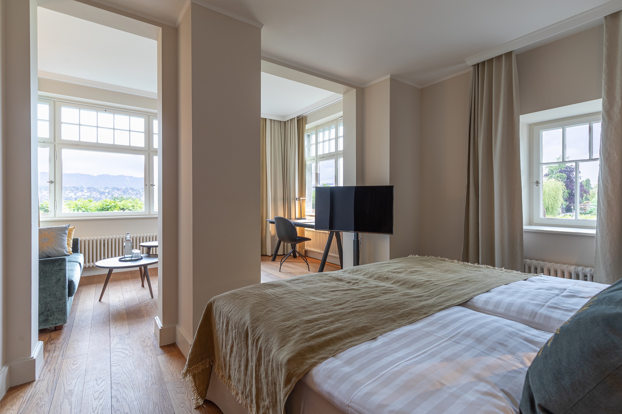 Helles, modernes Boutiquehotelzimmer mit Doppelbett, TV, Schreibtisch und Sitzecke. Große Fenster lassen natürliches Licht herein und bieten einen Blick ins Grüne. Helle Wände und Holzböden schaffen eine gemütliche Atmosphäre.
