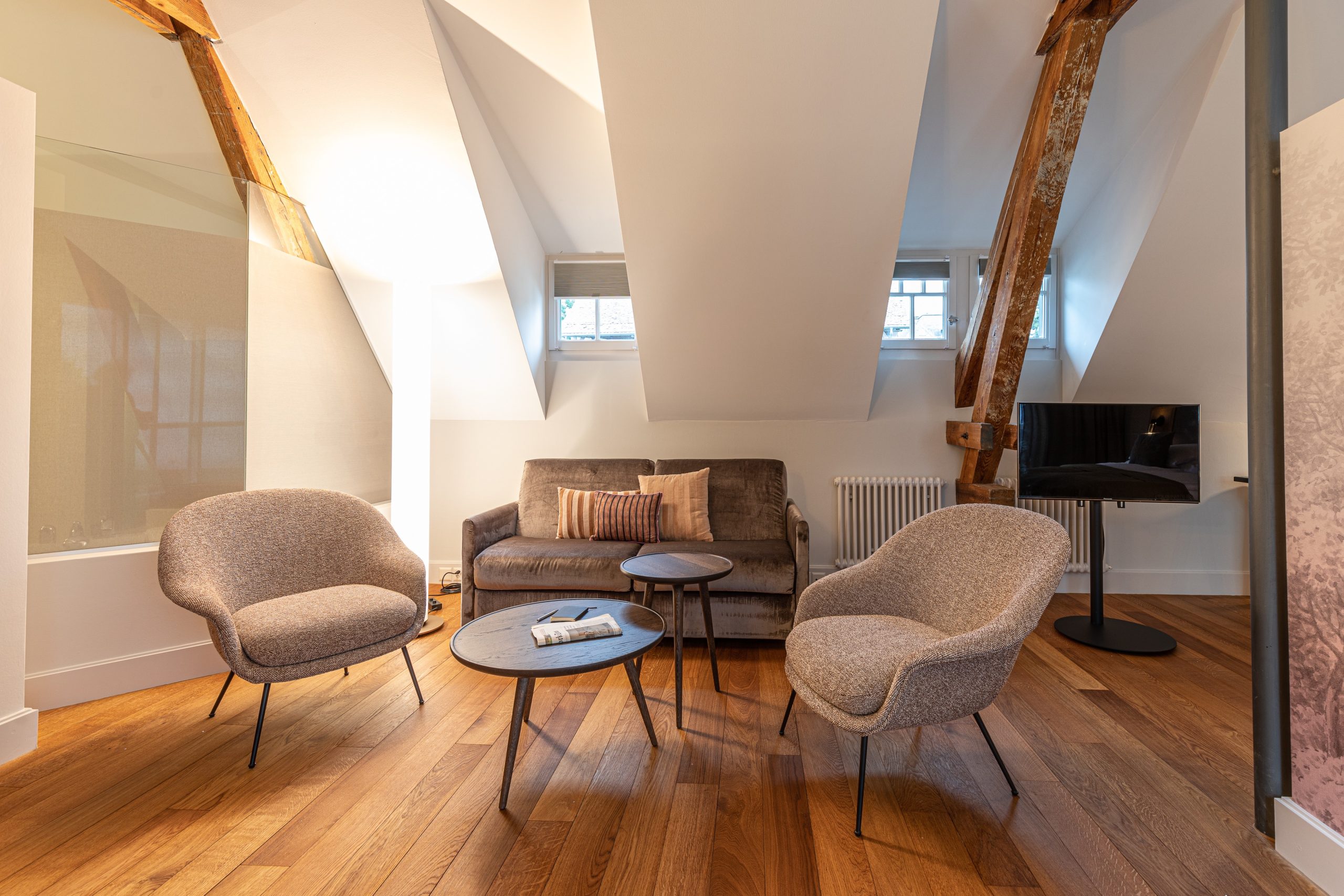 Modernes Dachgeschoss-Wohnzimmer mit schrägen Decken, grauen Sesseln im Stil eines Boutiquehotels, einem grauen Sofa und runden Couchtischen auf Holzfußboden. Freigelegte Balken und kleine Fenster verleihen dem gemütlichen Raum Charakter.