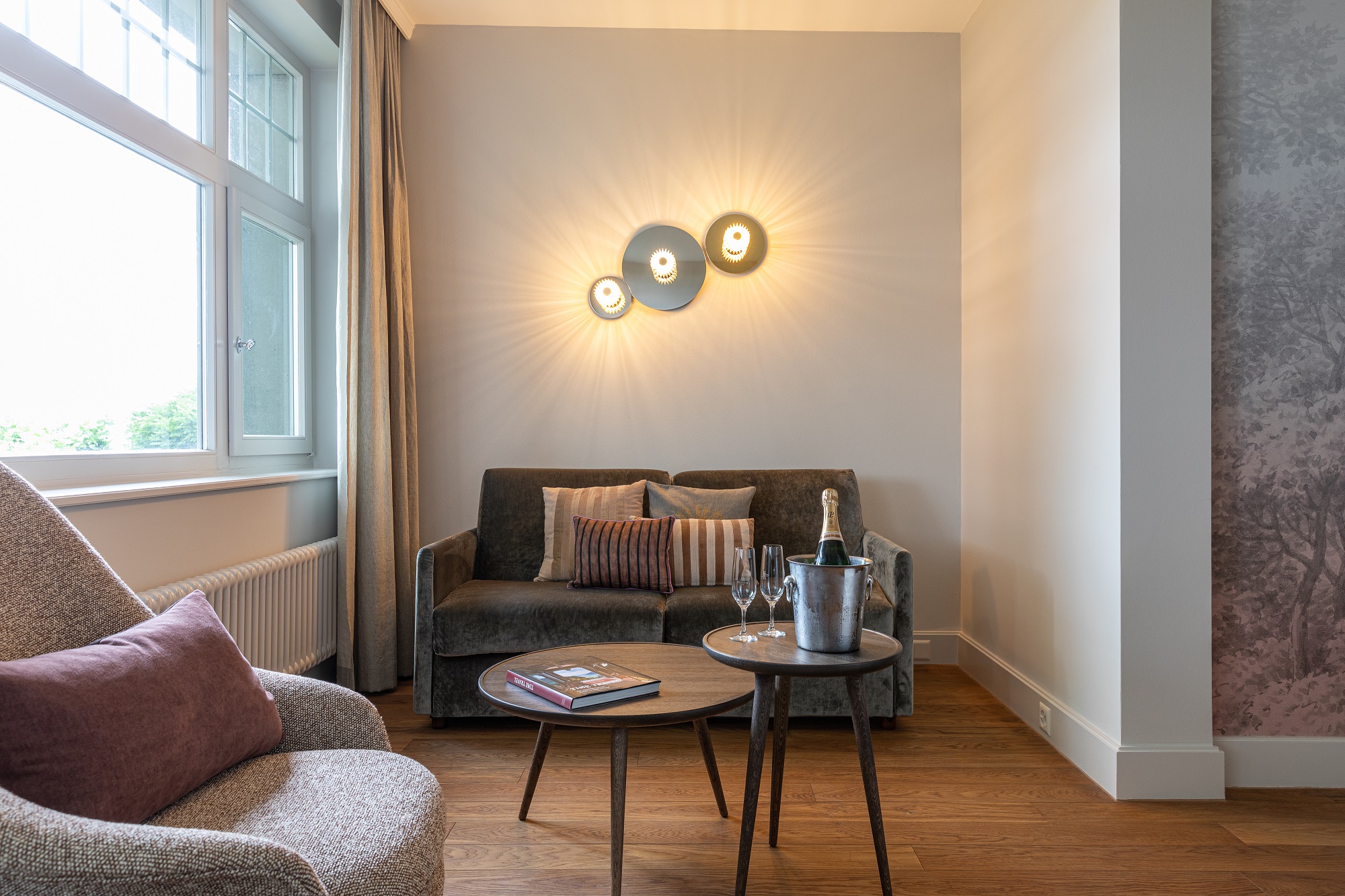 Ein modernes Wohnzimmer mit einem grauen Sofa, einem Sessel und runden Holztischen versprüht den Charme eines Boutiquehotels. Eine Champagnerflasche, zwei Gläser und Bücher schmücken die Tische. Wandleuchten schaffen eine warme Atmosphäre, während natürliches Licht durch ein offenes Fenster fällt.