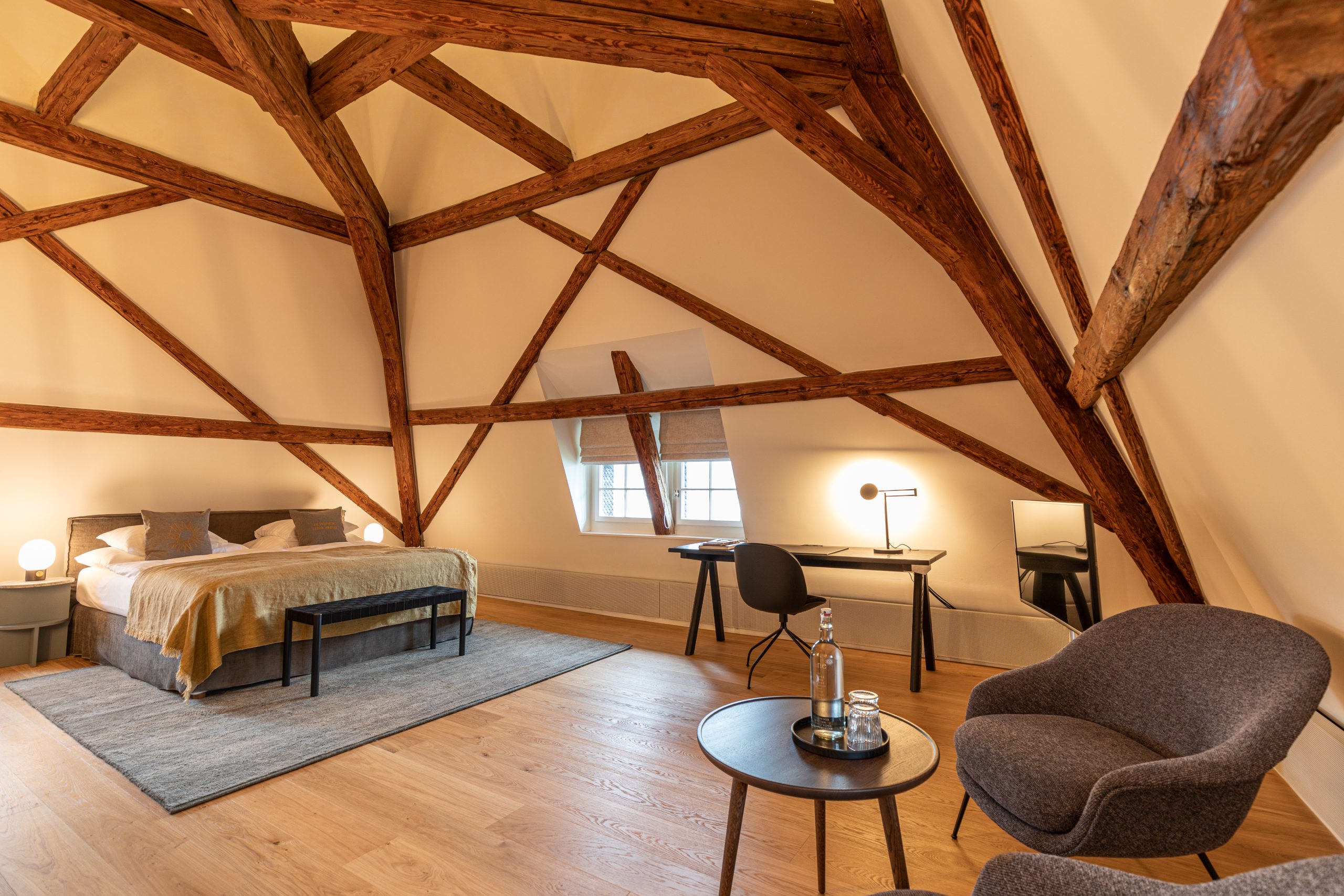 Geräumiges Boutiquehotel-Zimmer mit freiliegenden Holzbalken, einem großen Bett mit neutraler Bettwäsche, einem Schreibtisch und einer Lampe am Fenster sowie einer gemütlichen Sitzecke mit zwei Stühlen und einem runden Tisch auf Holzboden.