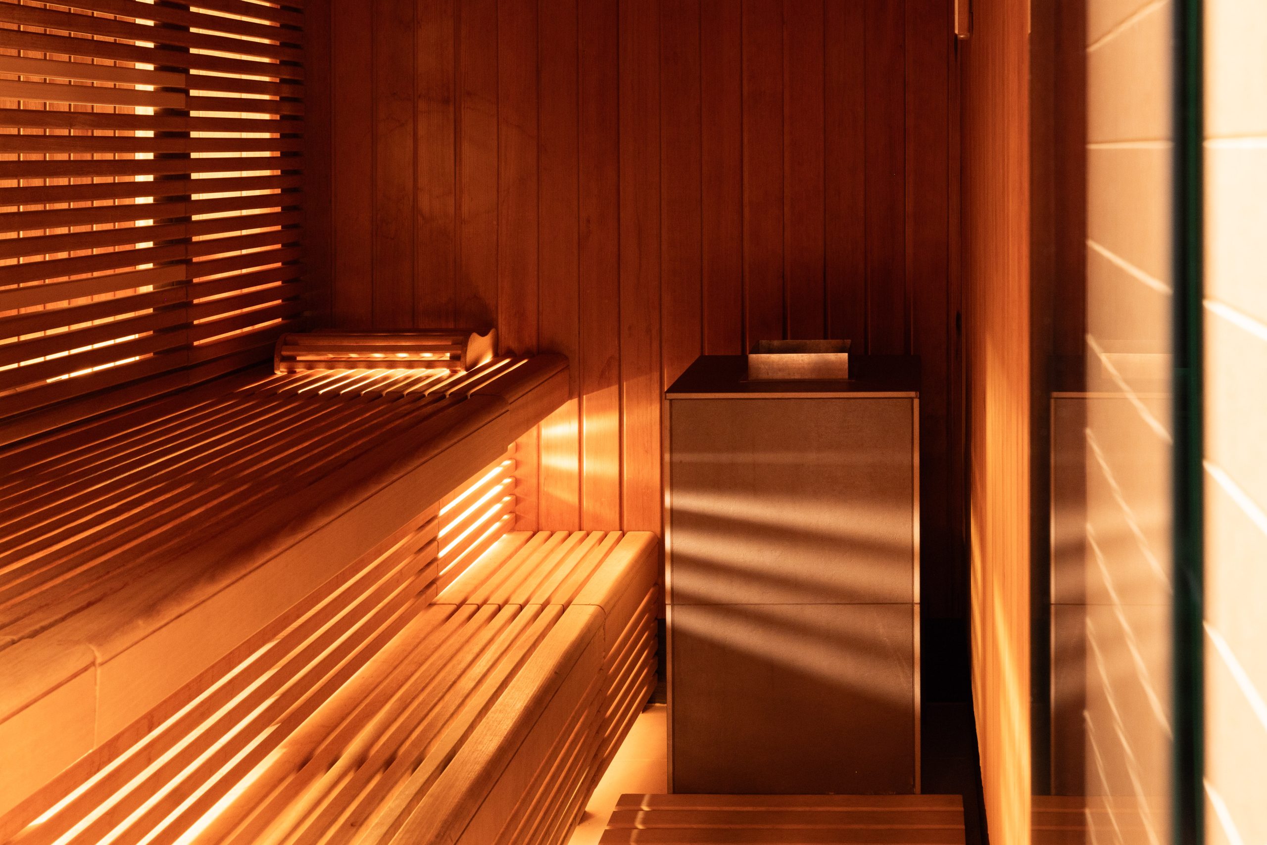 Eine Holzsauna im Boutiquehotel, in der das Sonnenlicht durch vergitterte Fenster fällt und gestreifte Schatten auf Bänke und Wände wirft. In der Ecke steht ein Ofen mit Steinen, der eine warme und einladende Atmosphäre schafft.