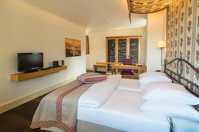 Ein modernes Boutiquehotelzimmer mit einem Doppelbett, gestreiften und gemusterten Kissen, einem an der Wand montierten Fernseher, einem Schreibtisch mit Stuhl und dekorativen Wandpaneelen. Der Raum verfügt über eine sanfte Beleuchtung und warme, neutrale Farben für einen gemütlichen Aufenthalt.