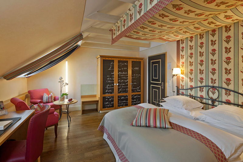 Gemütliches Boutiquehotelzimmer mit Doppelbett, gemustertem Baldachin und Tapete, gestreiften Kissen, Holzboden, Schreibtisch, roten Stühlen, kleinem runden Tisch und einem Kleiderschrank mit Schriftzug an den Türen.