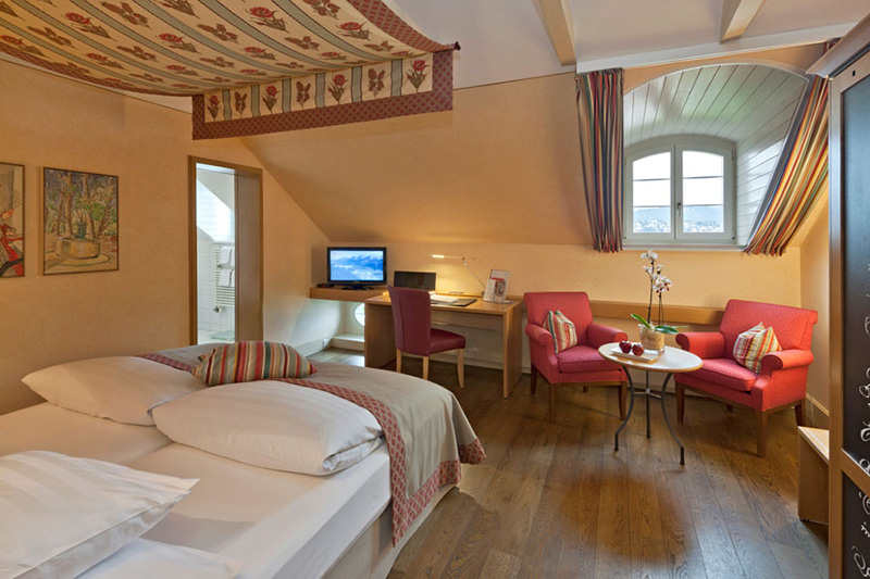 Gemütliches Boutiquehotelzimmer mit einem Doppelbett, zwei roten Sesseln um einen kleinen Tisch, einem Schreibtisch mit Computer, Holzboden, schräger Decke und einem Fenster mit gestreiften Vorhängen, das Tageslicht hereinlässt.