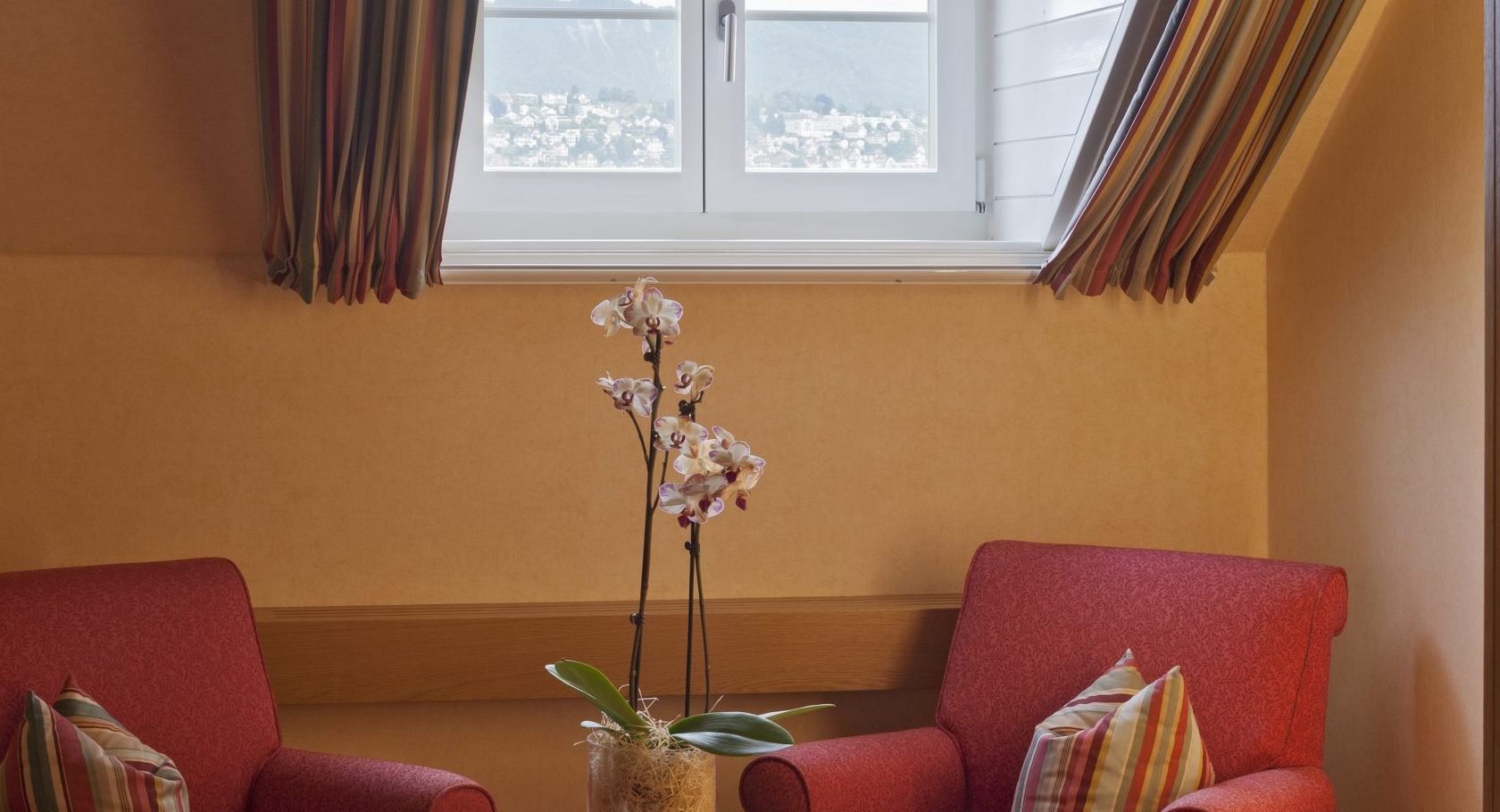 Zwei rote Sessel mit gestreiften Kissen sitzen um einen kleinen runden Tisch mit Äpfeln und einer Orchidee unter einem Bogenfenster in diesem Boutiquehotel, wo gestreifte Vorhänge einen herrlichen Blick auf die ferne Landschaft einrahmen.