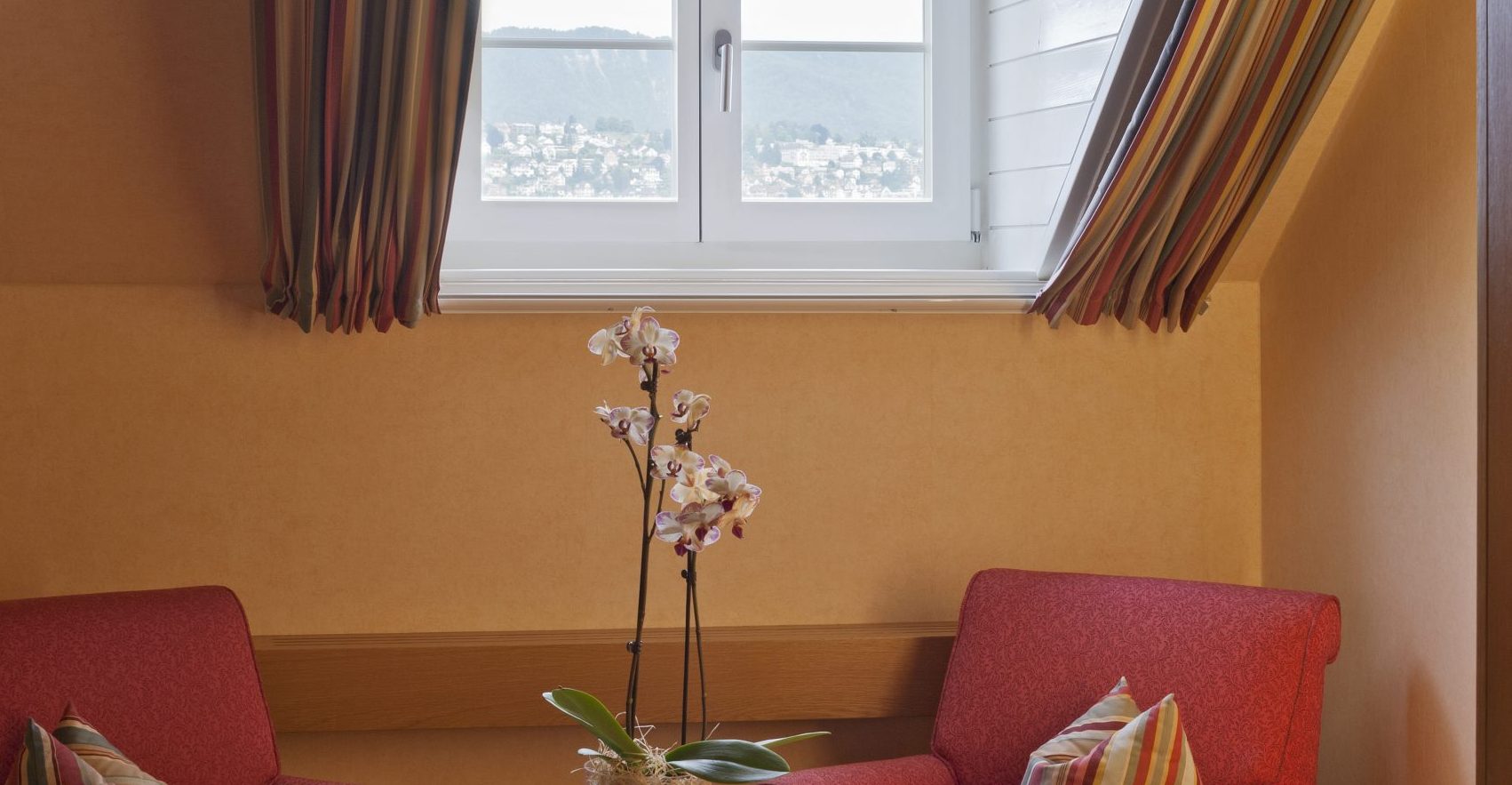 Zwei rote Sessel mit gestreiften Kissen sitzen um einen kleinen runden Tisch mit Äpfeln und einer Orchidee unter einem Bogenfenster in diesem Boutiquehotel, wo gestreifte Vorhänge einen herrlichen Blick auf die ferne Landschaft einrahmen.