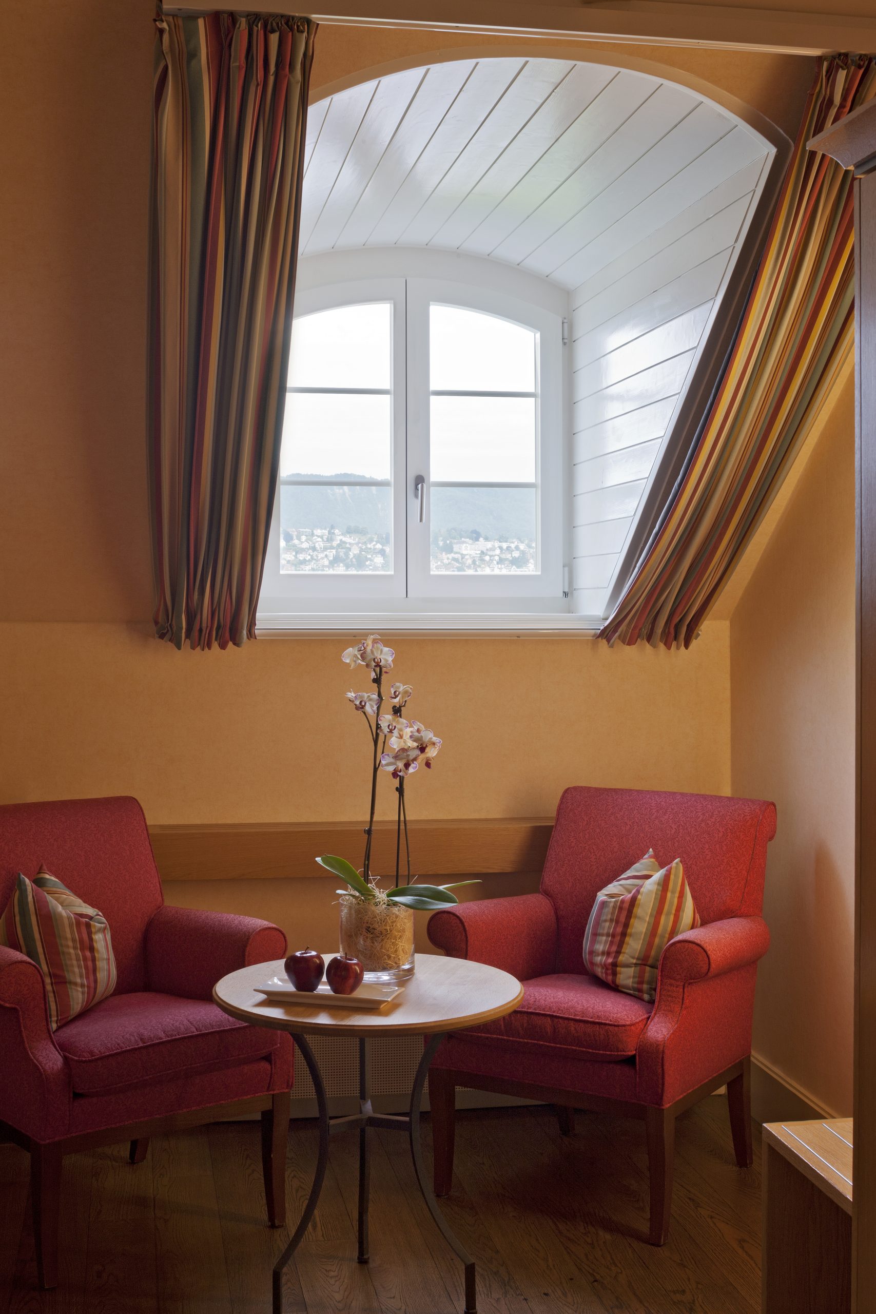 Zwei rote Sessel mit gestreiften Kissen sitzen um einen kleinen runden Tisch mit Äpfeln und einer Orchidee unter einem Bogenfenster in diesem Boutiquehotel, wo gestreifte Vorhänge einen herrlichen Blick auf die ferne Landschaft einrahmen.