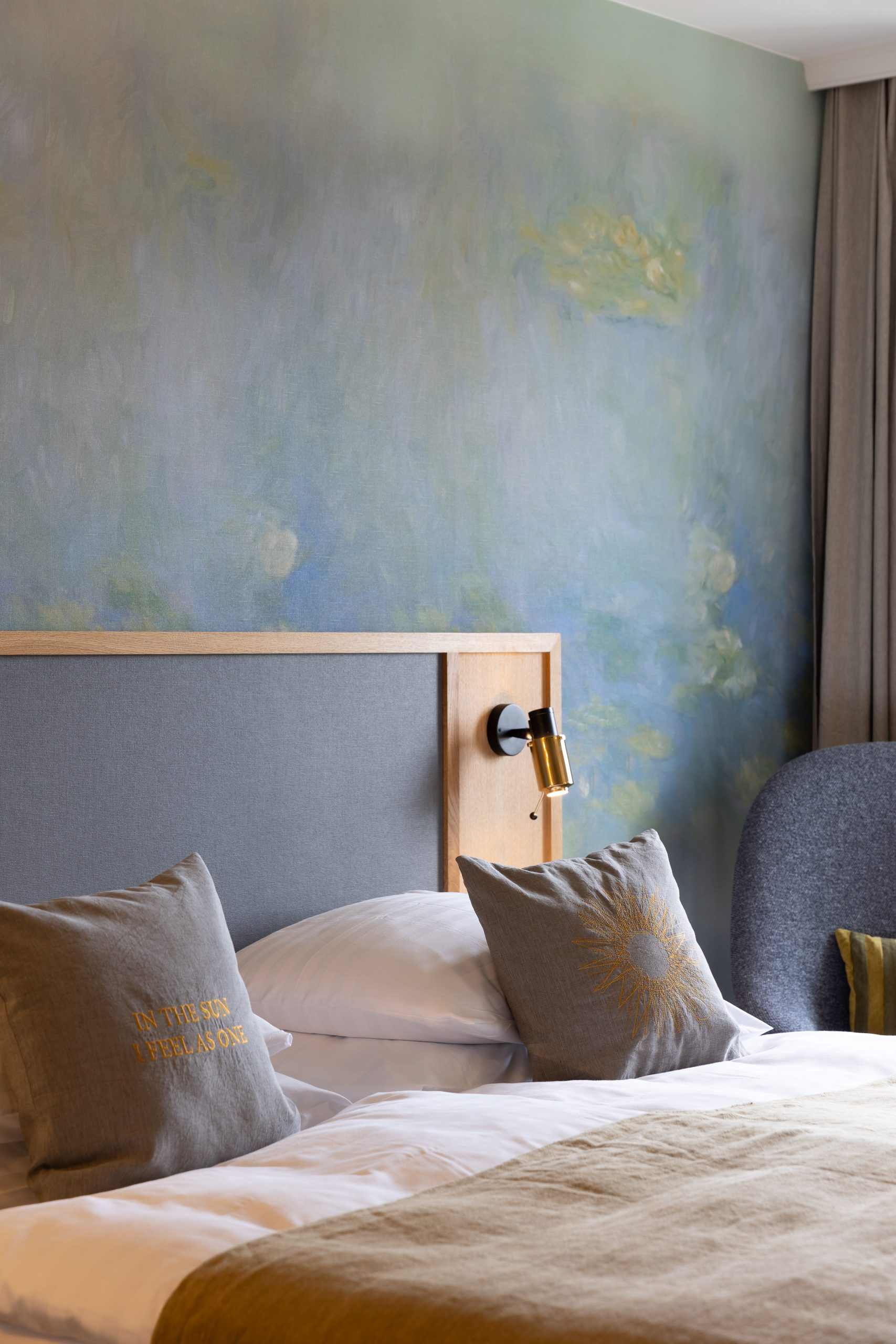 Ein ordentlich gemachtes Bett mit grauen und goldenen Akzentkissen, ein hölzernes Kopfteil, eine Leselampe und ein Wandgemälde in sanften Blau- und Grüntönen schaffen eine Boutiquehotel-Atmosphäre neben einem Fenster mit beigen Vorhängen.