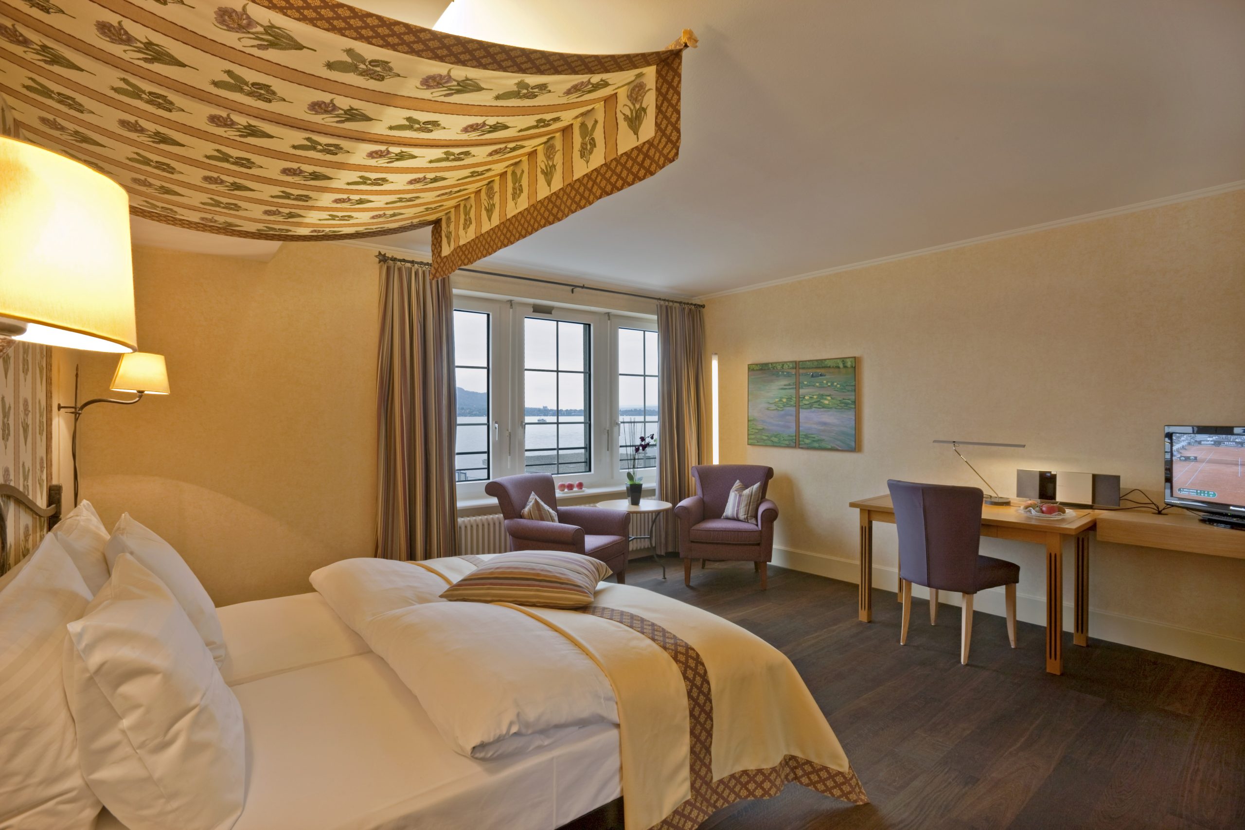 A cozy Deluxe Room Lake at Boutiquehotel Sonne features a double bed with canopy, two armchairs by large windows overlooking the water, a desk with lamp, and TV on a wooden table. The decor is warm and inviting.