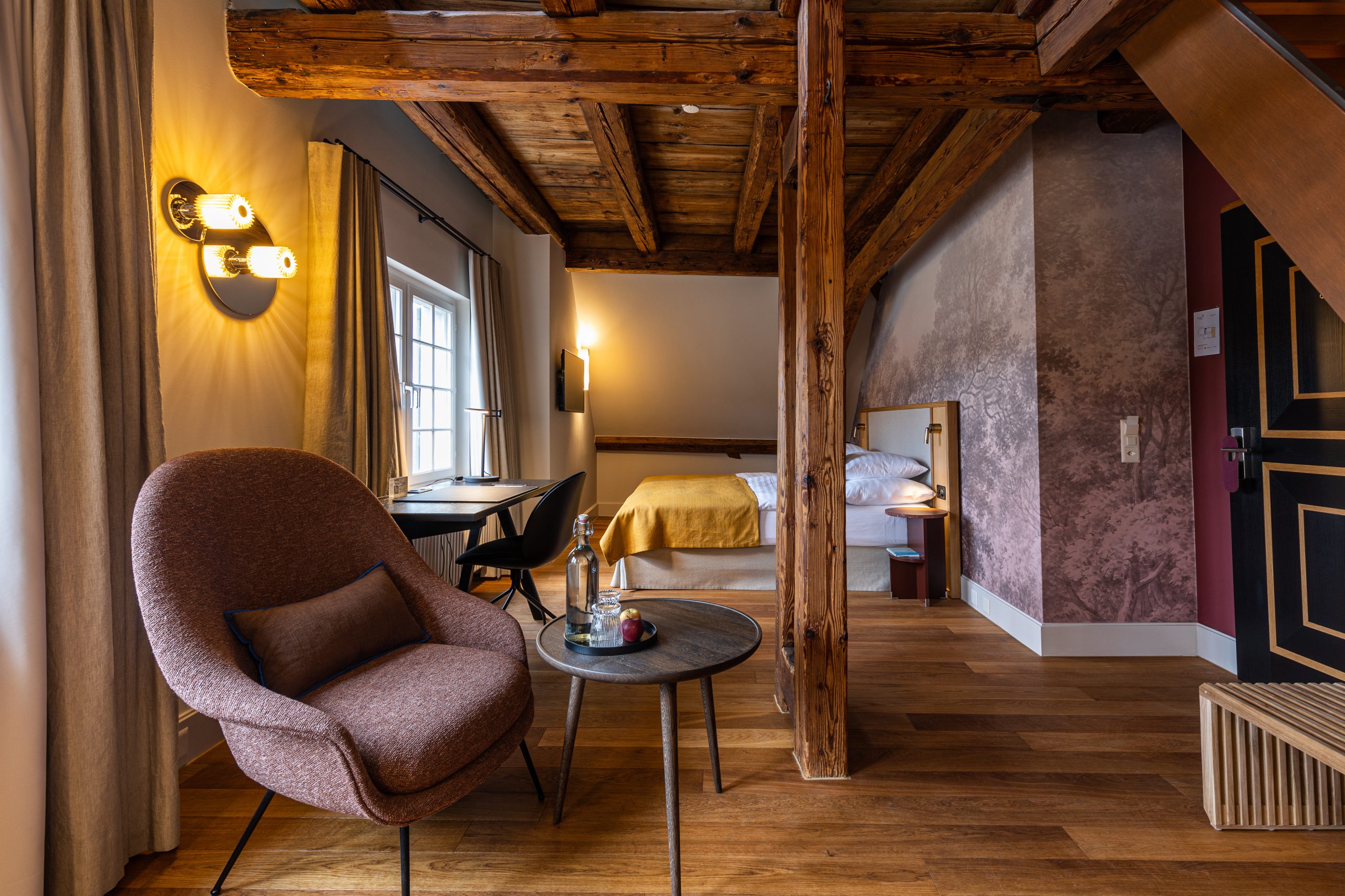 Gemütliches, modernes Familienzimmer mit Seesicht, freiliegenden Holzbalken, einem Sessel und Tisch im Vordergrund, einem Schreibtisch am Fenster und einem ordentlich gemachten Bett mit gelber Bettwäsche. Die warme Beleuchtung unterstreicht die einladende Atmosphäre.