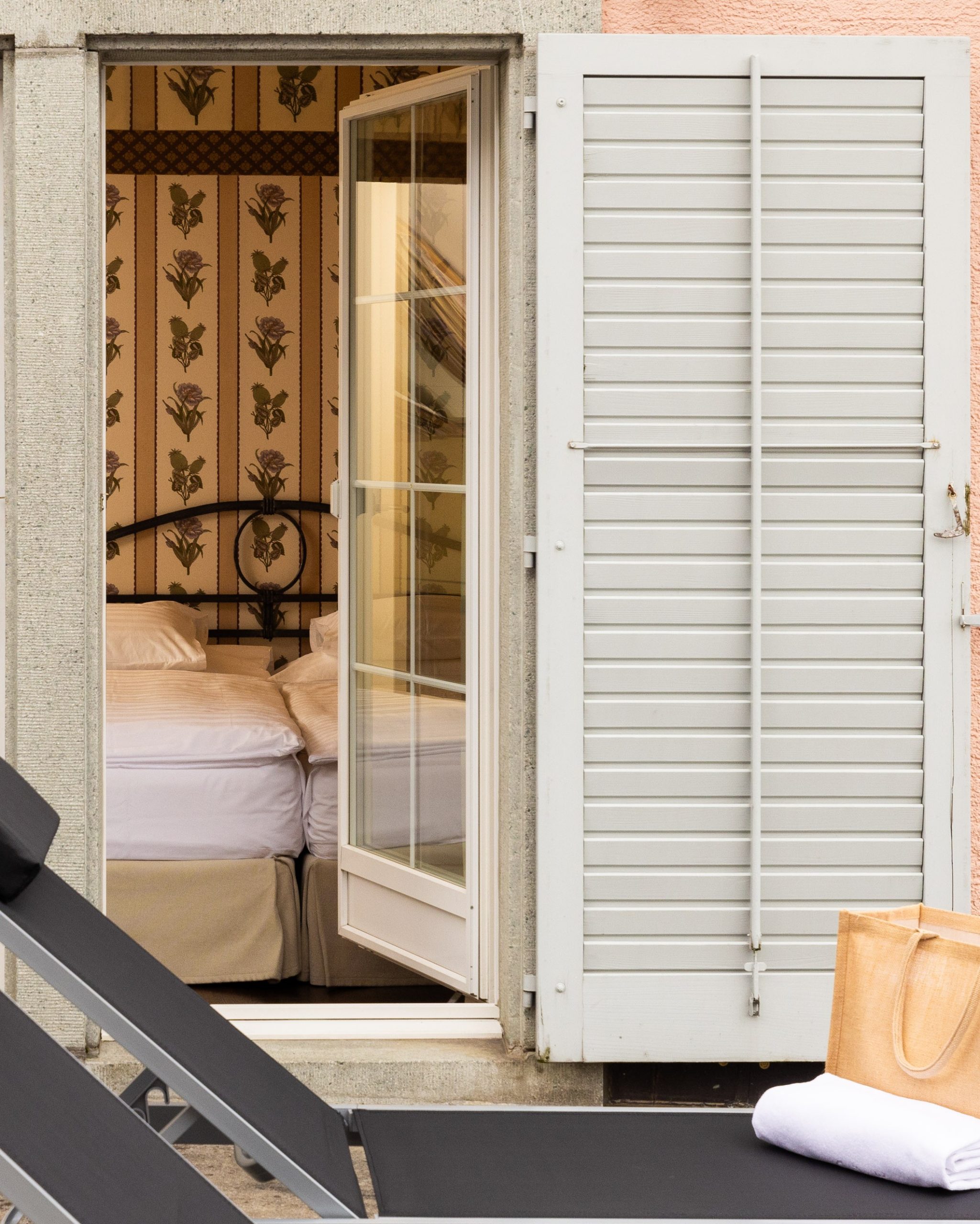 Ein gemütliches Deluxe-Zimmer mit Terrasse See verfügt über ein Doppelbett und eine Blumentapete, die durch offene Glastüren mit weißen Fensterläden sichtbar ist. Draußen auf der Terrasse erwarten Sie zwei schwarze Liegestühle und eine beige Tasche mit Handtüchern.