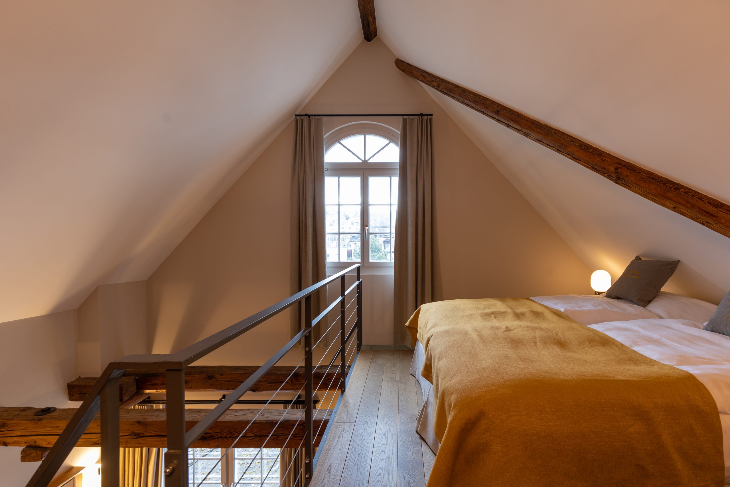 Ein gemütliches Familienzimmer mit Seesicht im Dachgeschoss, mit Dachschrägen, freiliegenden Holzbalken, einem Doppelbett mit senfgelber Bettwäsche, einer kleinen Lampe und einem Fenster mit Vorhängen, die Tageslicht hereinlassen.