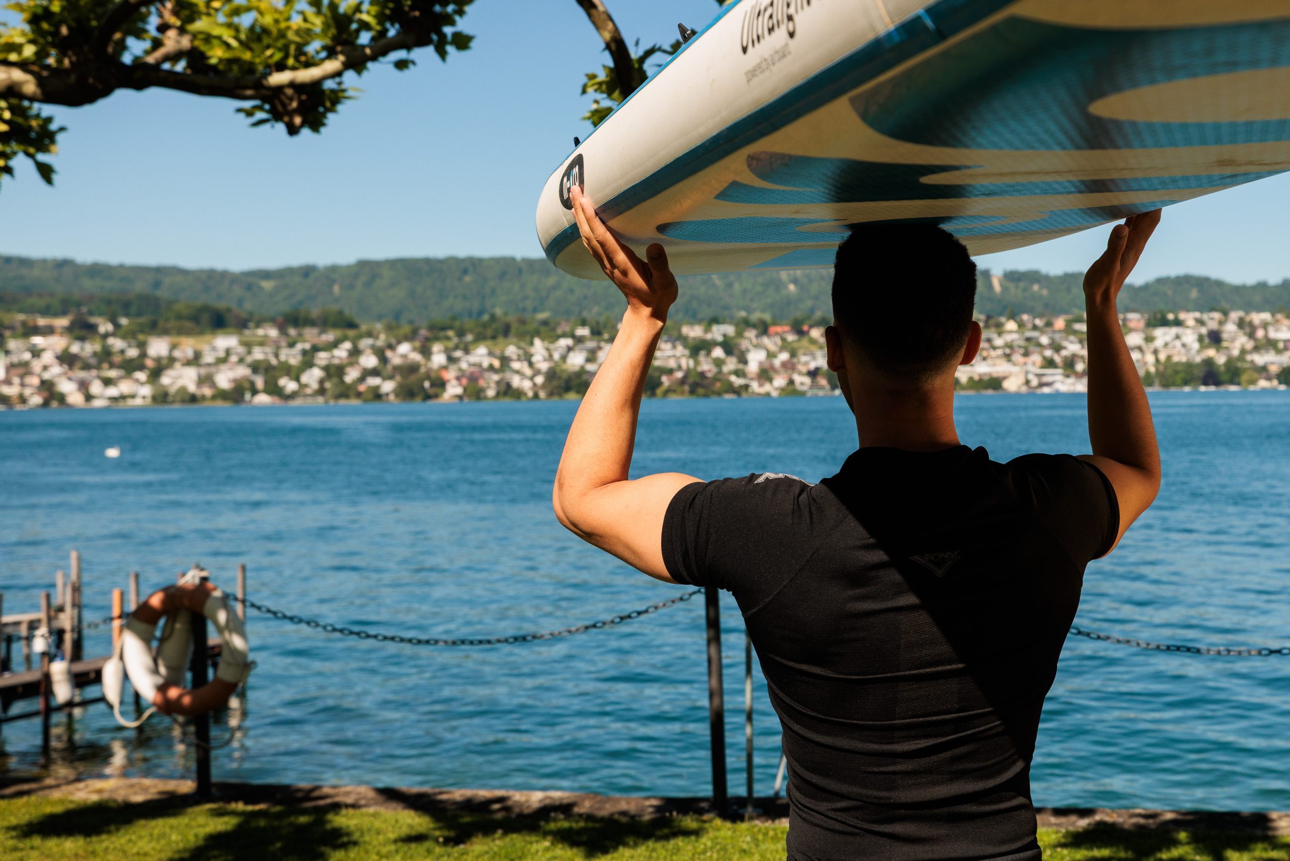 Eine Person steht an einem Seeufer und hält ein Paddelbrett auf dem Kopf, mit Blick auf das Wasser. In der Nähe befindet sich eine Anlegestelle mit einem Rettungsring, und im Hintergrund ist eine Stadt mit Hügeln unter einem klaren Himmel zu sehen.