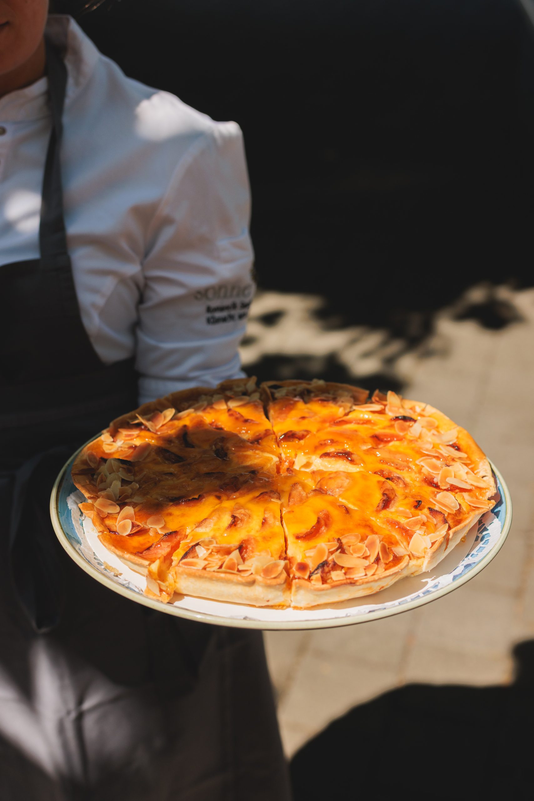 A person wearing a white shirt and gray apron holds a plate with a sliced fruit tart topped with peach slices and almond flakes.