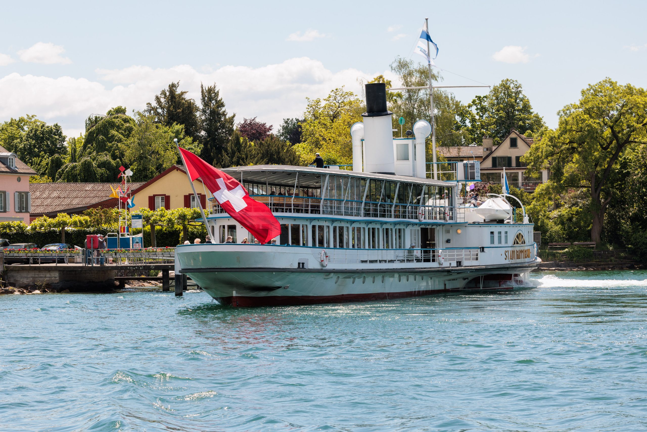 Ein altes Dampfschiff mit einer großen Schweizer Flagge segelt auf einem See in Ufernähe, mit Häusern, Bäumen und einem blauen Himmel mit Wolken im Hintergrund.