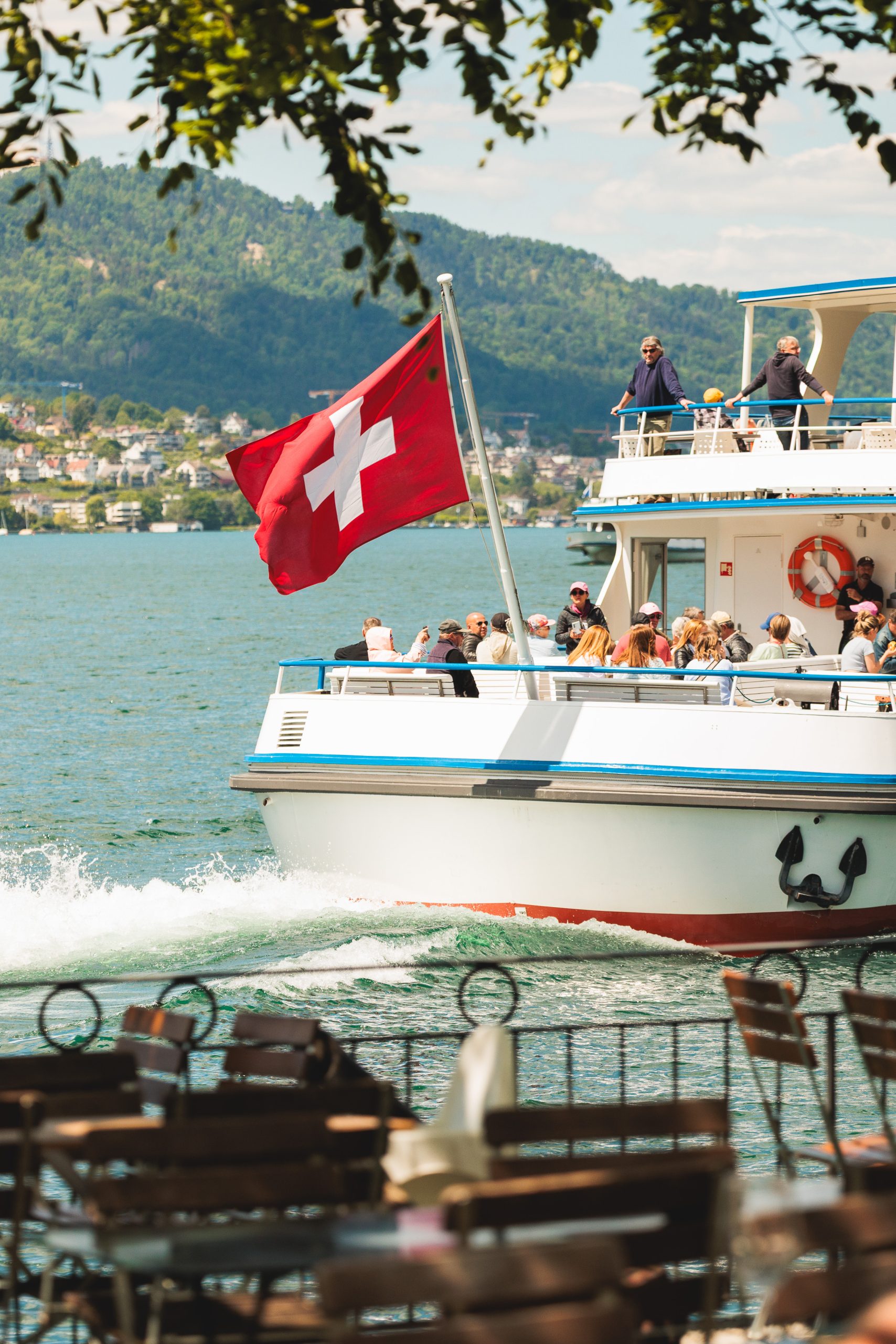 Ein Boot mit einer großen Schweizer Flagge segelt auf einem See und trägt Menschen auf seinem Deck. Im Vordergrund sind leere Cafétische zu sehen, im Hintergrund sind grüne Hügel und Gebäude am See zu sehen.