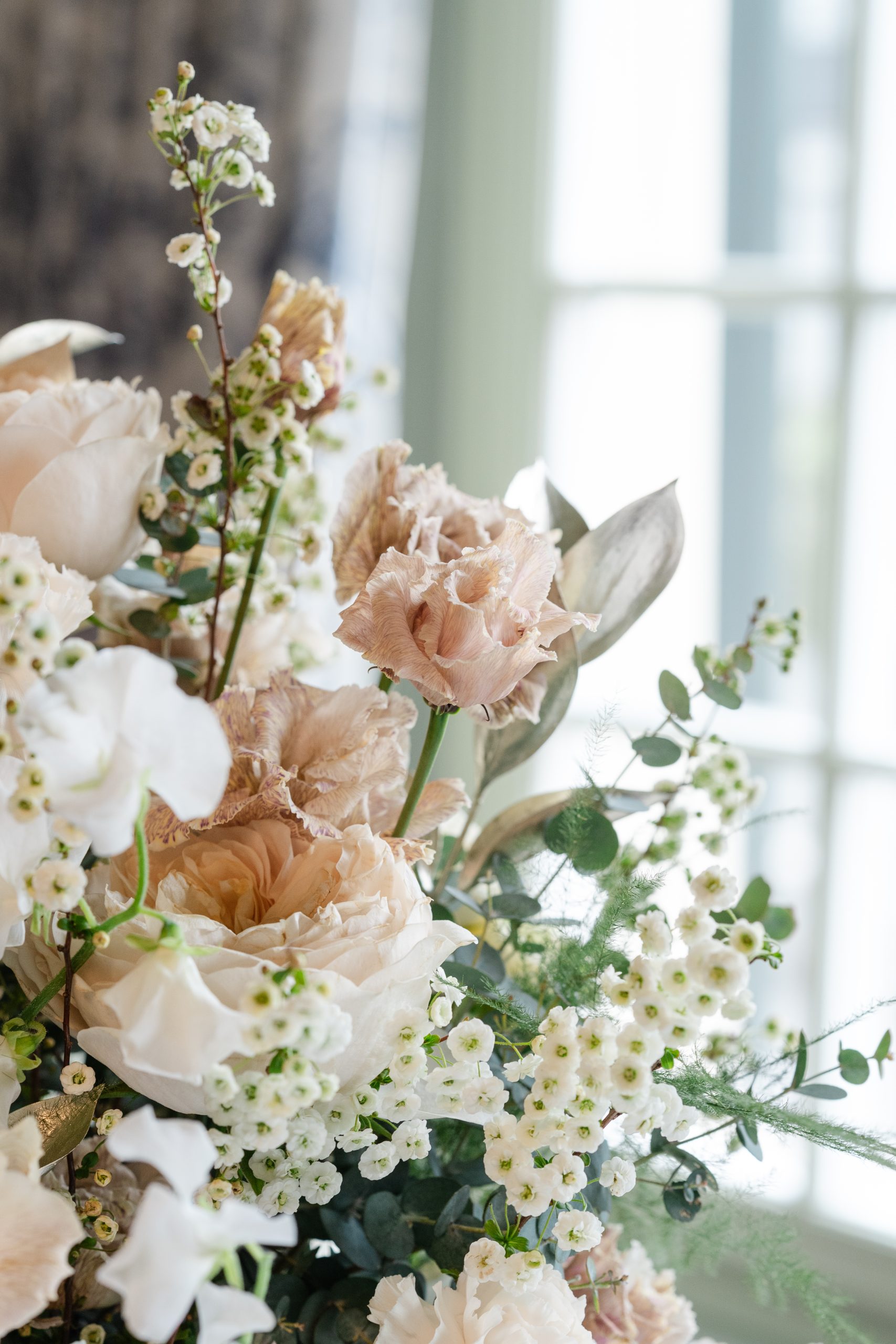 A close-up of a bouquet featuring soft pink roses, delicate white flowers, and greenery, with natural light coming through a window in the background.