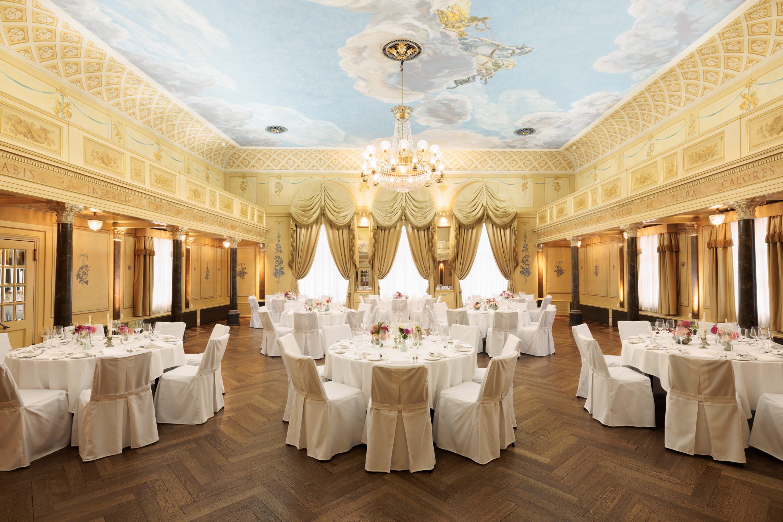 Ein eleganter Bankettsaal mit runden Tischen, die mit weißen Tüchern bedeckt und von weißen Stühlen umgeben sind. Die hohe Decke ist mit Wolken und Putten bemalt, und große Fenster mit goldenen Vorhängen säumen die Wände. In der Mitte hängt ein Kronleuchter.