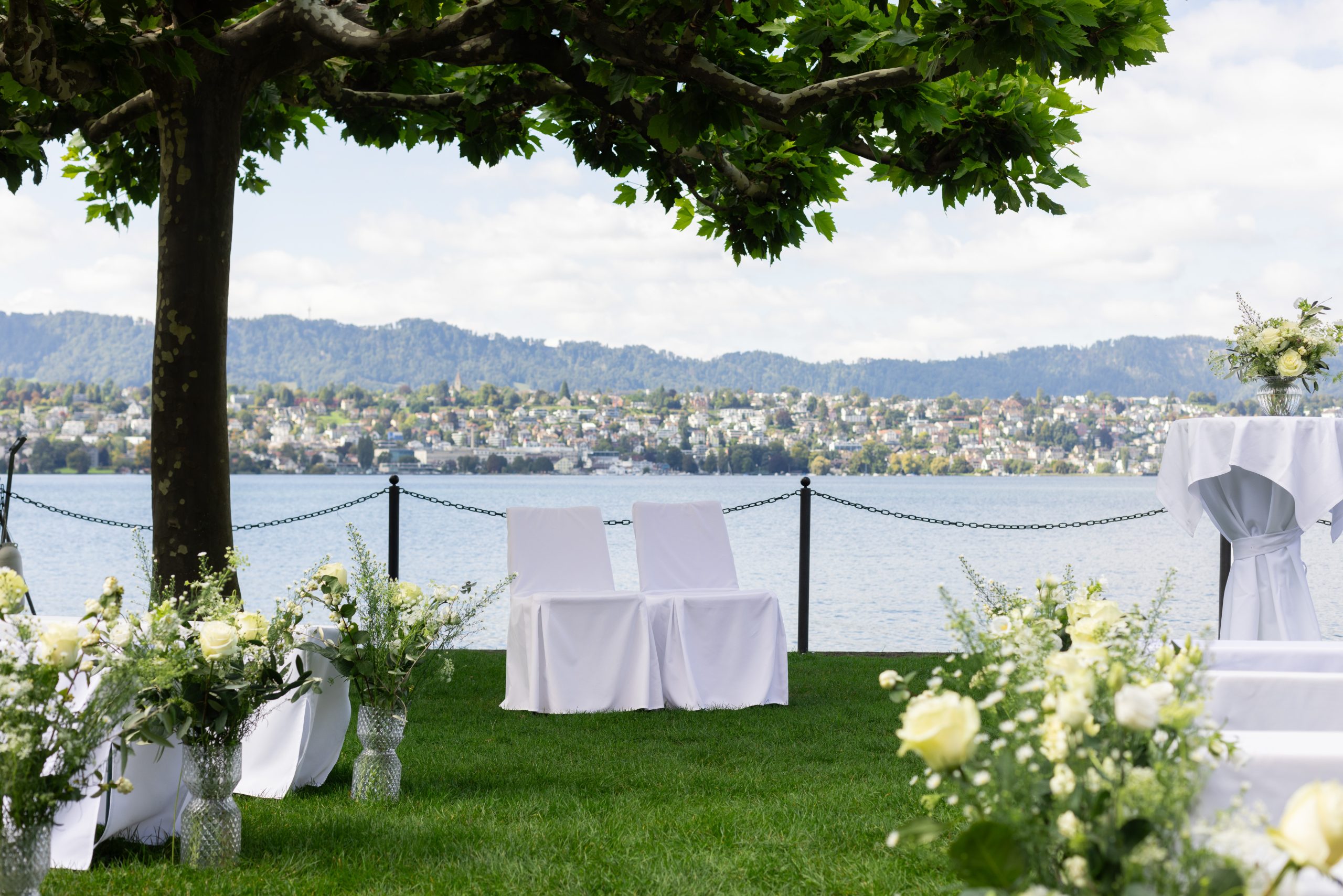 Hochzeit im Freien an einem See, mit weiß bezogenen Stühlen, Blumenarrangements und einem Tisch unter einem großen Baum. Ein malerischer Blick auf das Wasser und entfernte Häuser auf Hügeln im Hintergrund.