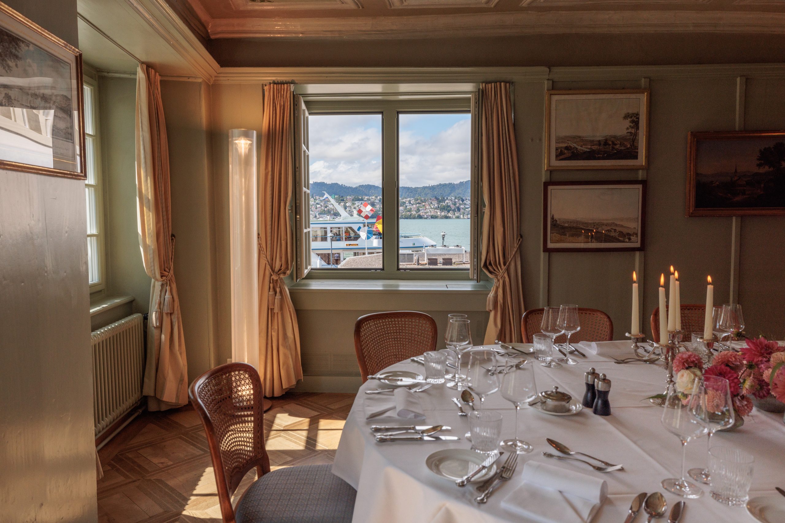 Im Speisesaal der Prunkstube des Event-Hotels Sonne steht ein runder, mit Blumen und Kerzen geschmückter Tisch mit Blick auf einen See mit Booten durch breite Fenster mit Vorhängen. Elegante Gemälde zieren die Wände.