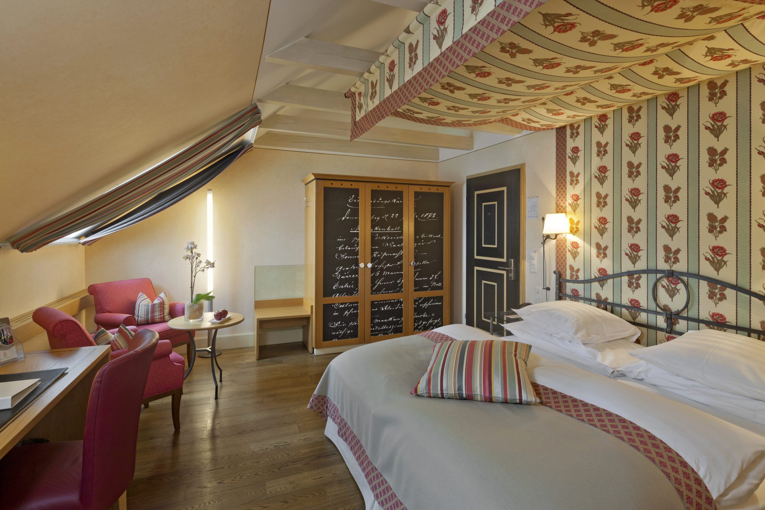 Das gemütliche Boutiquehotel Sonne Doppelzimmer See verfügt über ein Doppelbett, gestreiftes und geblümtes Dekor, einen kleinen Tisch mit roten Stühlen, einen hölzernen Kleiderschrank mit Schriftzug an den Türen, Holzböden und eine schräge Decke mit geblümten Tapetenakzenten.