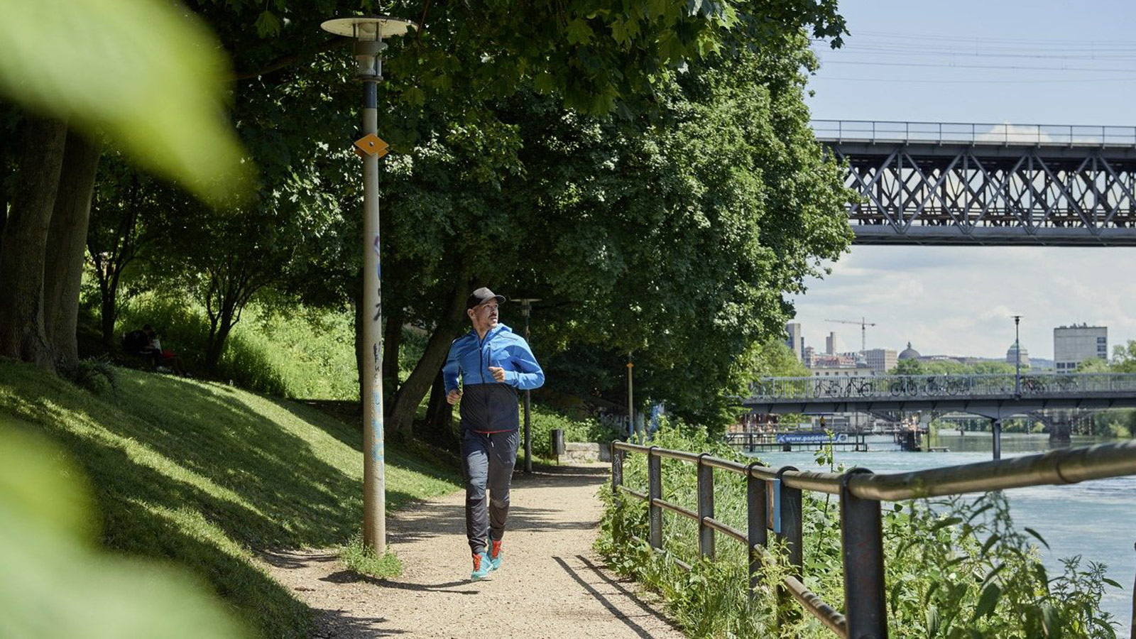 Eine Person joggt an einem sonnigen Tag einen von Bäumen gesäumten Uferweg entlang und genießt das örtliche Freizeitangebot, während im Hintergrund eine Metallbrücke und städtische Gebäude zu sehen sind.