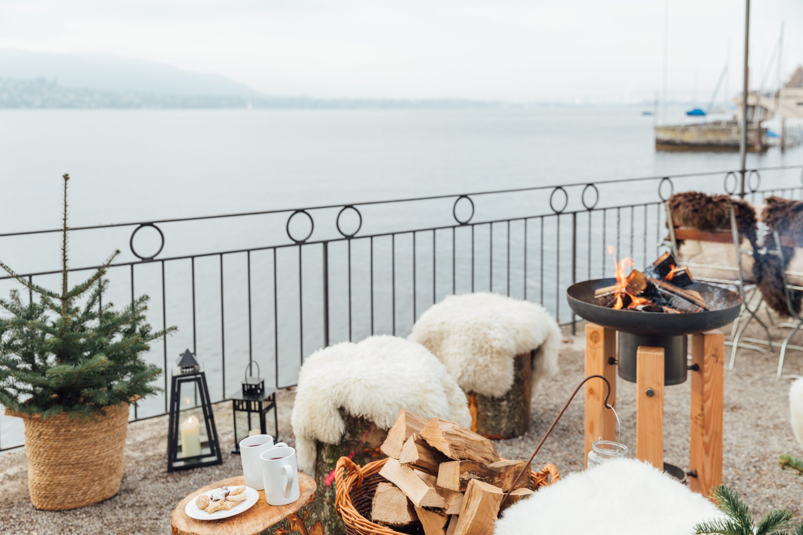 Eine gemütliche Seeterrasse im Restaurant Küsnacht mit Baumstumpfsitzen, die mit flauschigen weissen Decken bedeckt sind, einer brennenden Feuerstelle, Tassen und Keksen auf einem kleinen Tisch, einem Korb mit Feuerholz, einer Laterne und einer kleinen getopften Tanne.