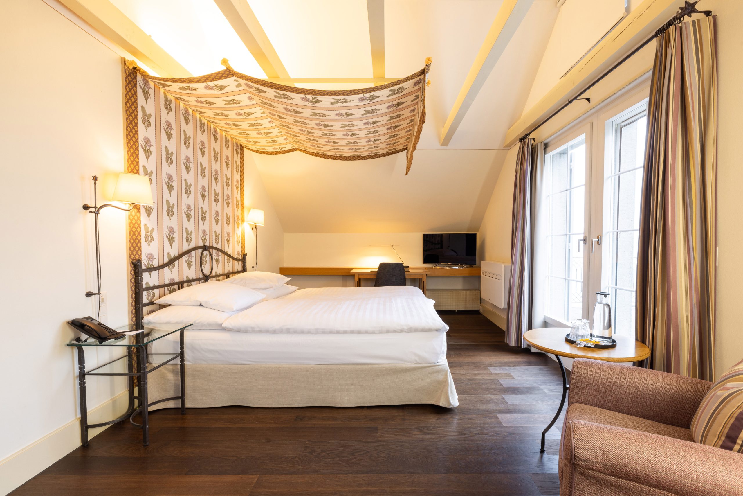 Ein gemütliches Hotelzimmer am Zürichsee mit einem Doppelbett unter einem dekorativen Baldachin, gestreiften Vorhängen, einem kleinen runden Tisch mit einem Teeservice, einem Sessel, einem Schreibtisch, einem Stuhl und einem Fernseher - alles in warmen, neutralen Tönen.