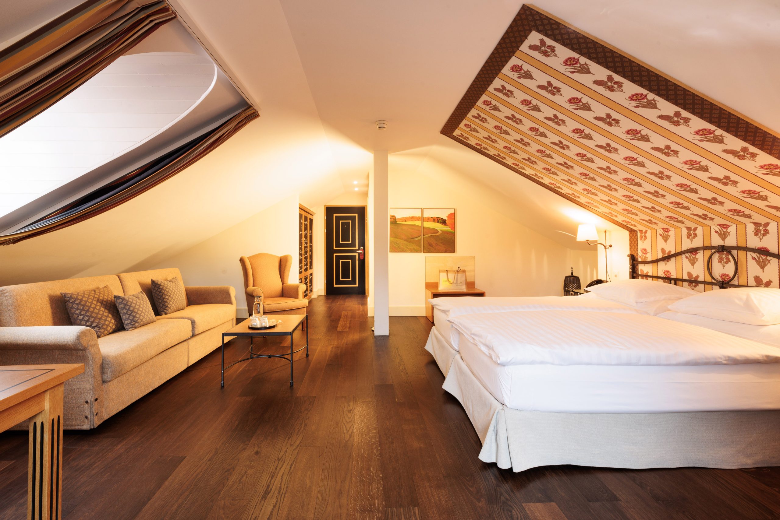 Ein gemütliches Hotelzimmer im Dachgeschoss am Zürichsee mit einem Doppelbett, einer gemusterten Schrägdecke, Holzböden, einem beigen Sofa, einem Sessel und einem kleinen Tisch mit einer Flasche und einem Glas. Die warme Beleuchtung schafft eine einladende Atmosphäre.