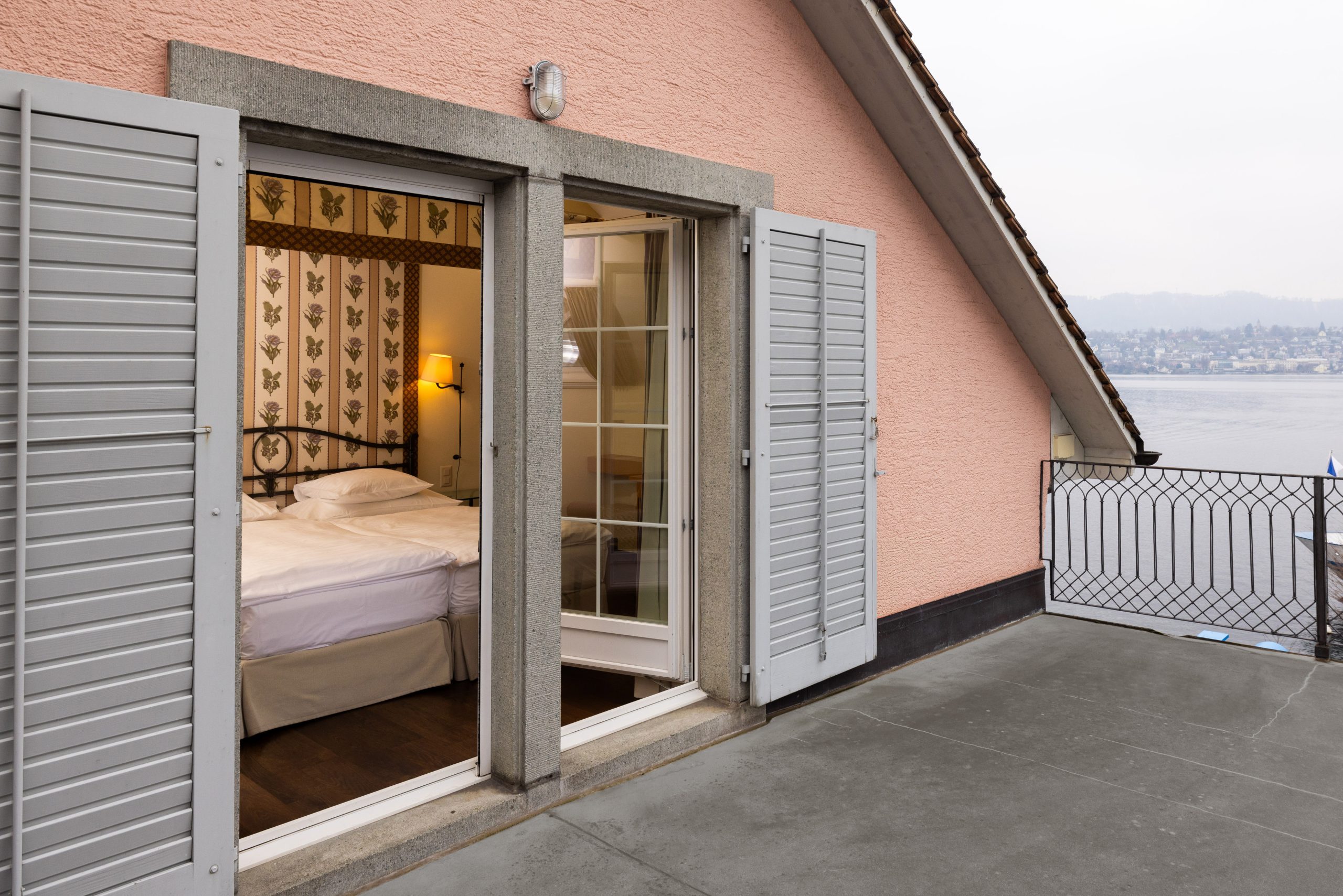 Ein Balkon mit grauen Fensterläden öffnet sich und gibt den Blick frei auf ein gemütliches Hotelzimmer am See mit einem Doppelbett, gemusterter Tapete und einer Lampe; der Balkon blickt auf ein Gewässer mit Hügeln in der Ferne.