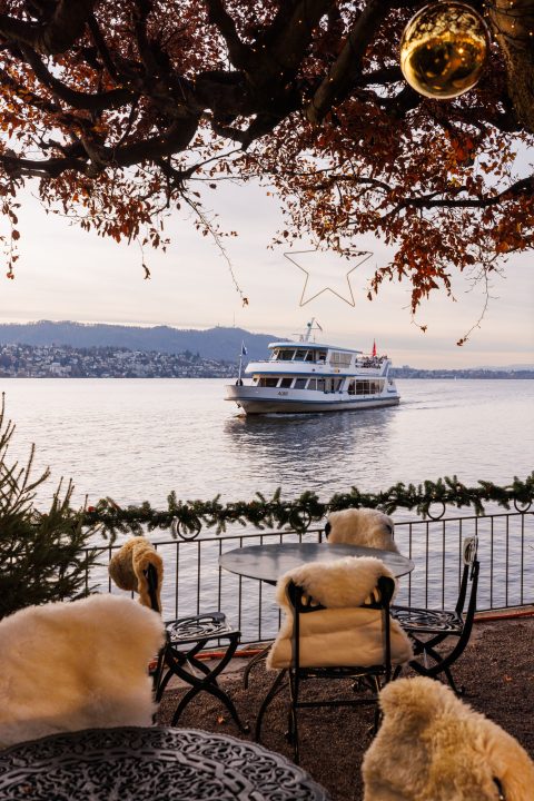 Ein gemütliches Café am See, das Chalet am See, mit fellbezogenen Stühlen und Blick auf ein Boot, das auf einem ruhigen See fährt. Herbstlaub umrahmt die Szene mit Weihnachtsdekorationen und einem sternförmigen Licht über dem Wasser. In der Ferne sind Hügel und Gebäude zu sehen.