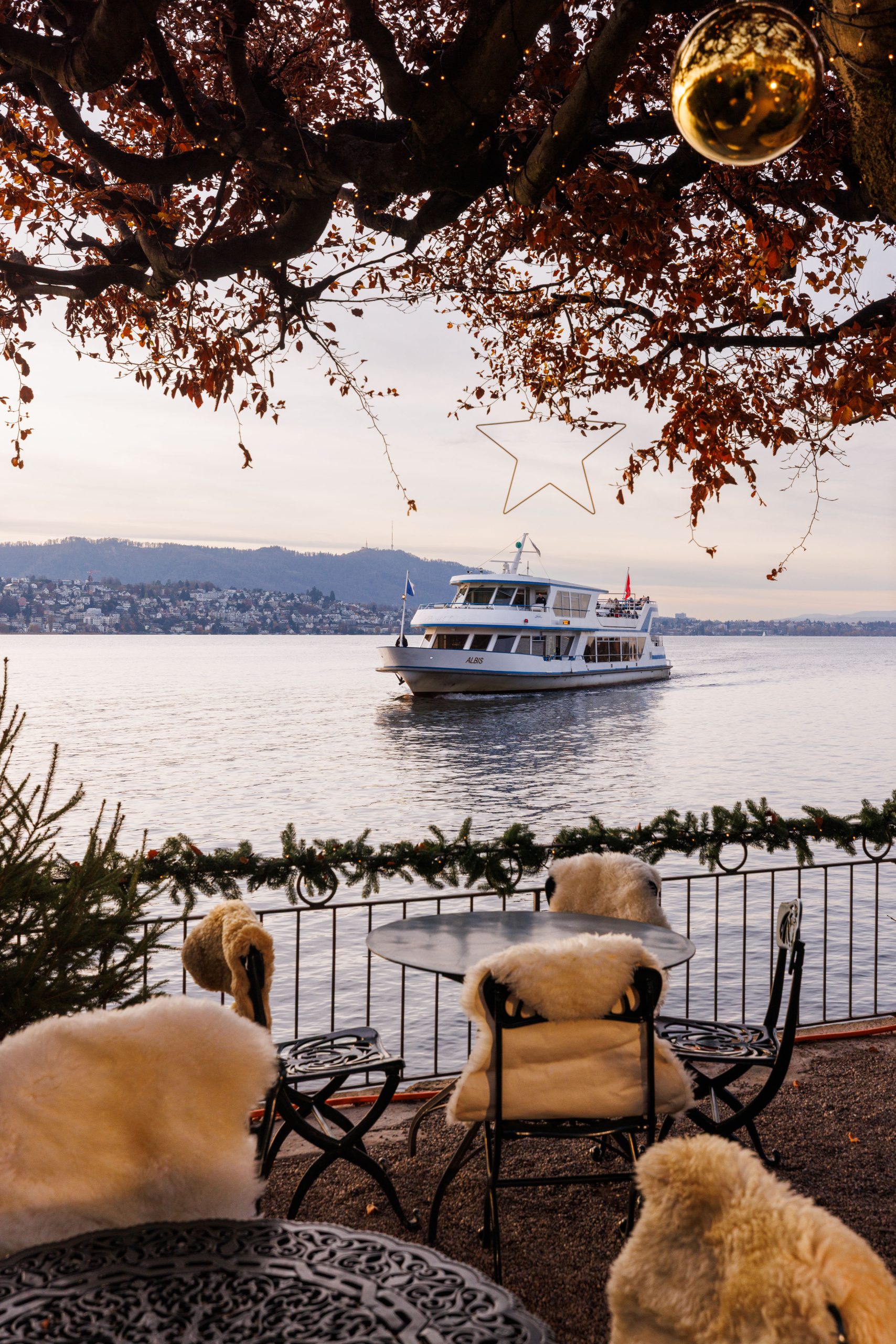 Ein gemütliches Café am See, das Chalet am See, mit fellbezogenen Sesseln blickt auf einen ruhigen See, auf dem ein Boot vorbeifährt. Herbstliche Bäume und festliche Dekorationen, darunter ein Stern und eine Girlande, umrahmen die Szene.