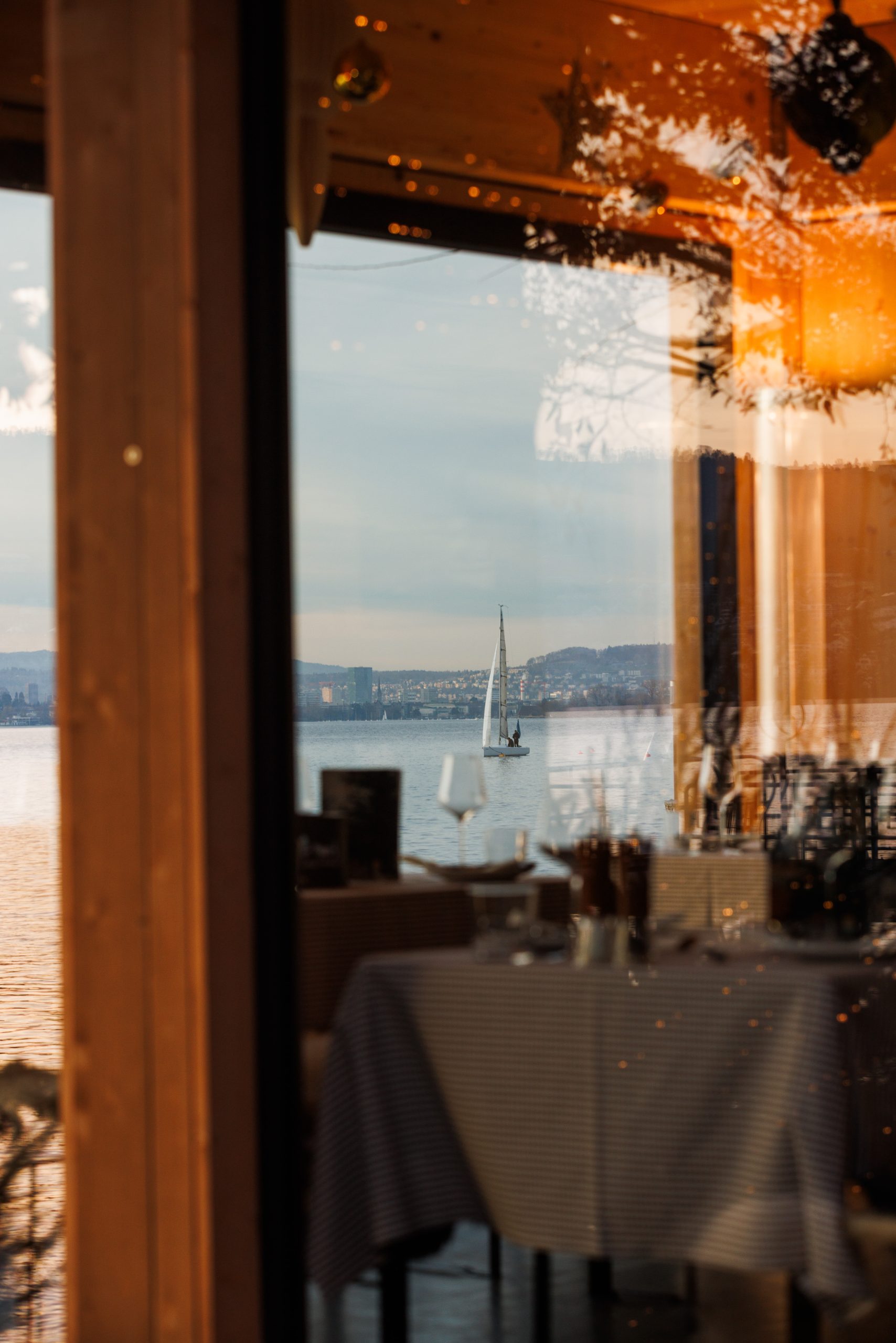 Ein Segelboot gleitet auf einem ruhigen See, gesehen durch ein Fenster eines gemütlichen Restaurants. Im Fenster spiegeln sich warmes Innenlicht und ein gedeckter Esstisch mit Gläsern und Geschirr, so dass Innen- und Außenszenen miteinander verschmelzen.