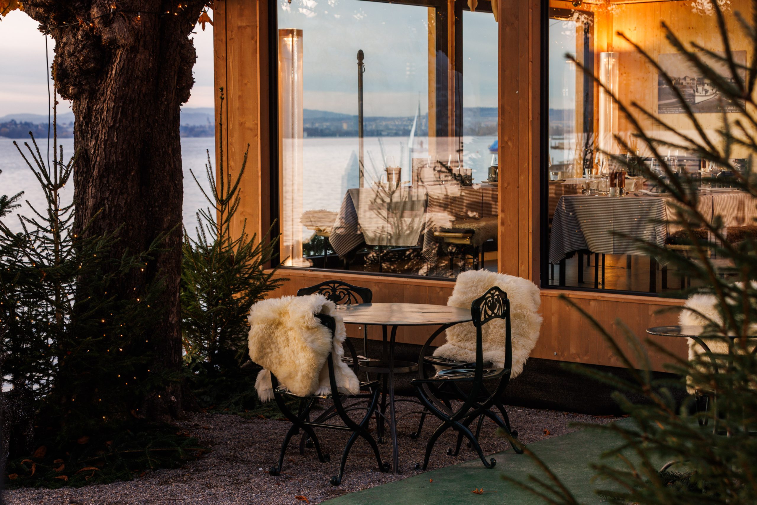 Das Außencafé im Chalet am See verfügt über zwei mit weißem Pelz bespannte Stühle, einen runden Tisch und einen herrlichen Blick auf den See. In den großen Fenstern spiegelt sich das Wasser, während Pinienbäume und sanfte Beleuchtung eine gemütliche, einladende Atmosphäre schaffen.