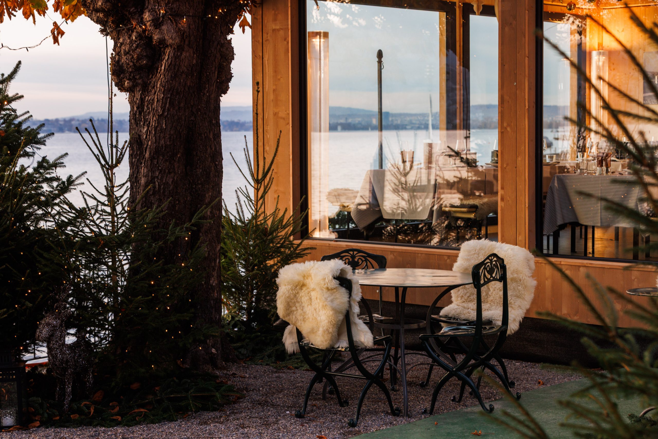 A cozy outdoor seating area with two chairs draped in white fur around a small table, surrounded by evergreens beside a wooden chalet by the lake, overlooking calm waters at sunset.
