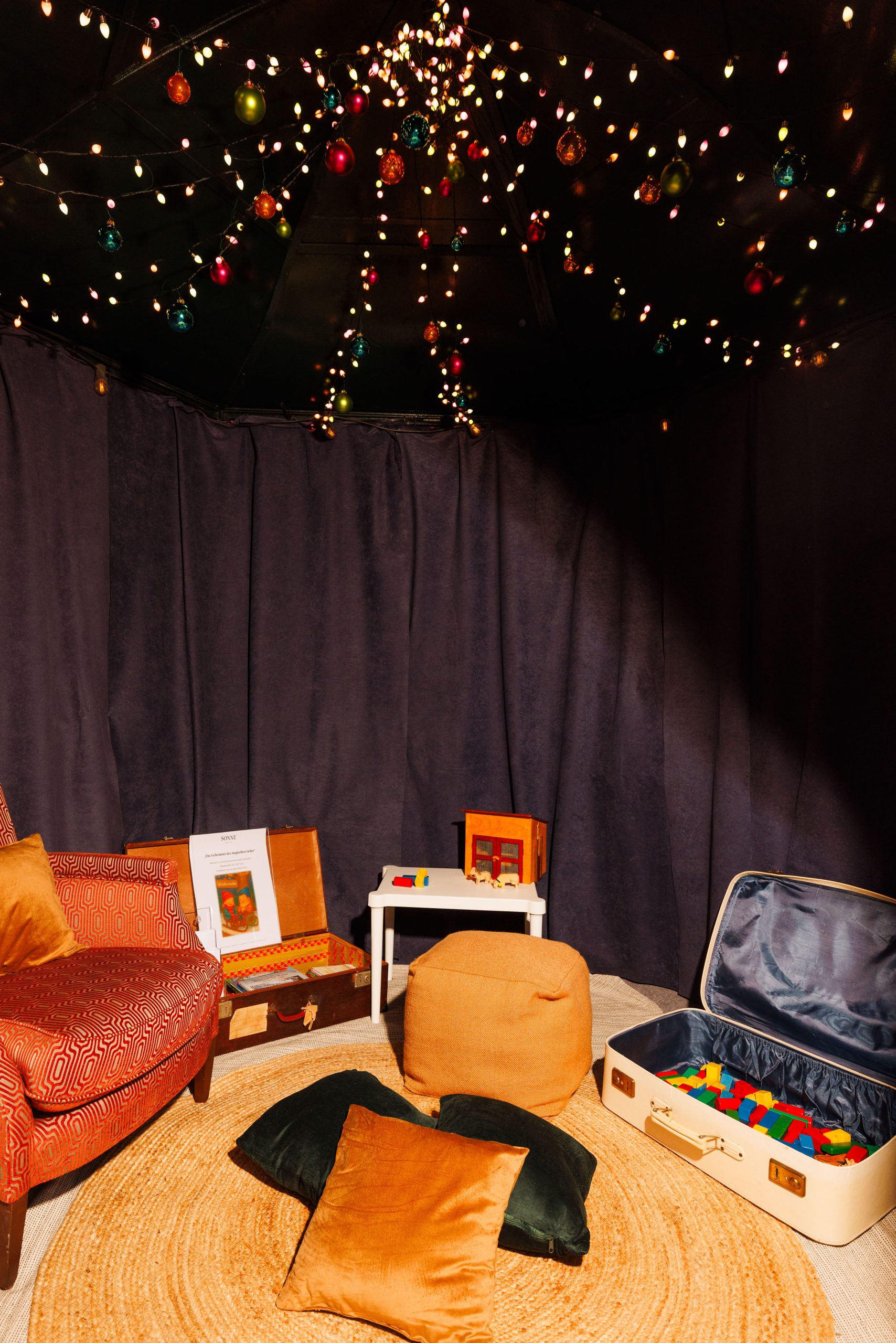 Gemütliches Spielzimmer mit dunklen Vorhängen, warmer Beleuchtung und Lichterketten mit bunten Ornamenten an der Decke. Der Raum enthält einen gemusterten Sessel, einen Hocker, Kissen, einen runden Teppich, einen Koffer mit Spielzeug und einen kleinen weißen Tisch mit Stühlen.
