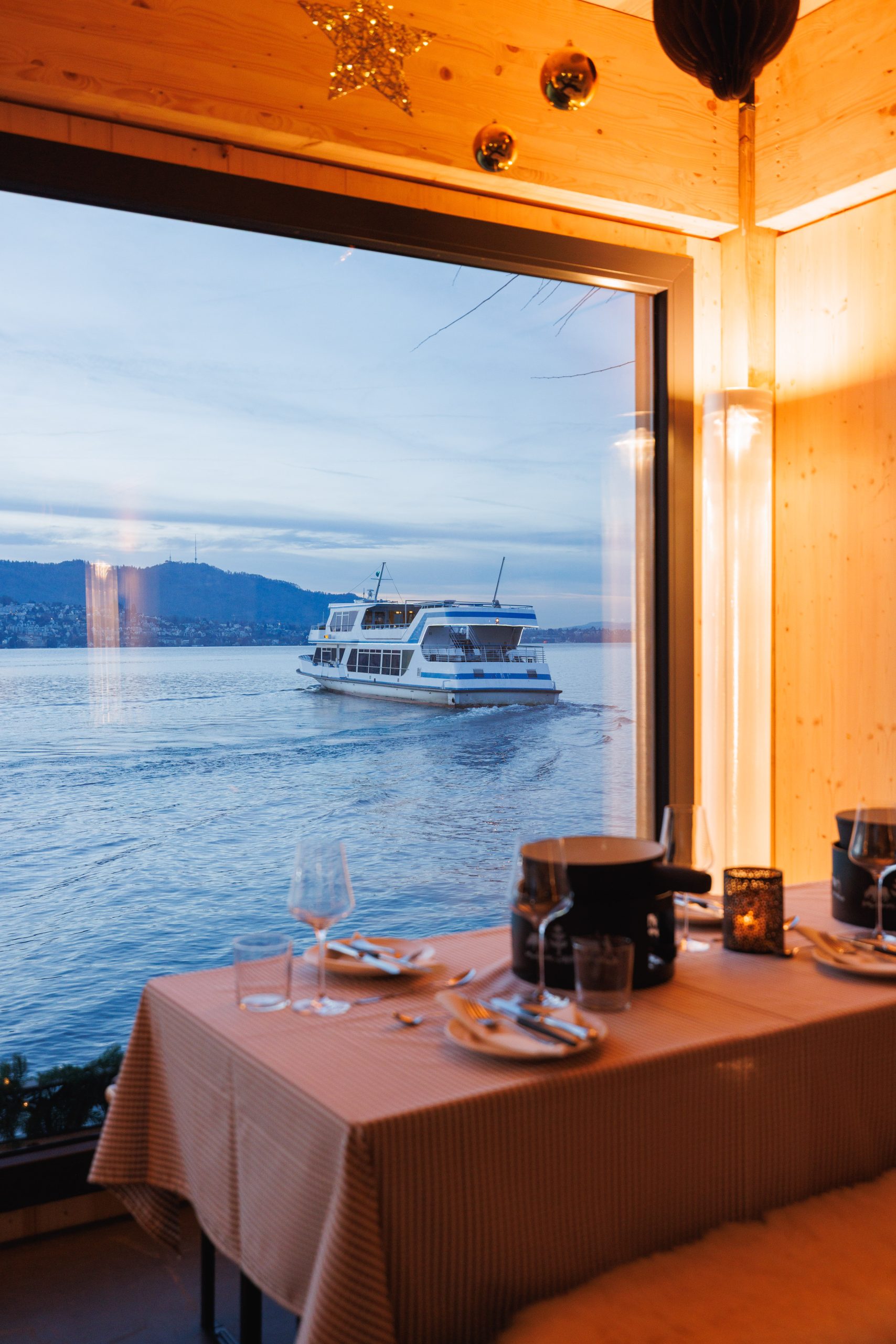 Ein gemütlicher Esstisch für zwei Personen mit Fondue blickt auf ein großes Fenster, durch das ein Boot auf einem ruhigen See bei Sonnenuntergang segelt, mit fernen Hügeln unter einem blauen Himmel.
