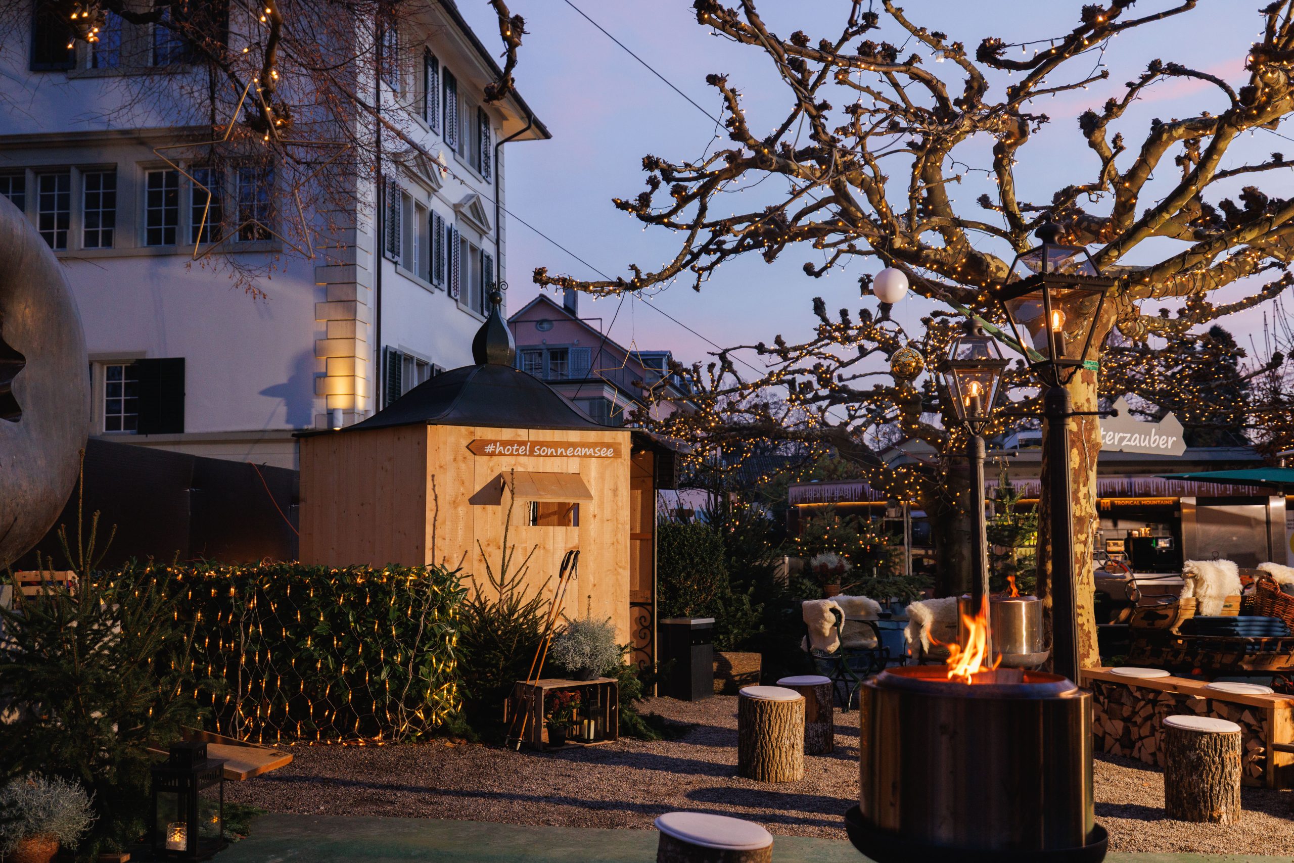 Ein gemütlicher Sitzbereich im Freien mit einer Feuerstelle, einem Baum mit Lichterketten, rustikalen Holzbänken und einer kleinen Holzhütte in der Nähe eines großen Gebäudes schafft in der Dämmerung eine warme und einladende Atmosphäre.